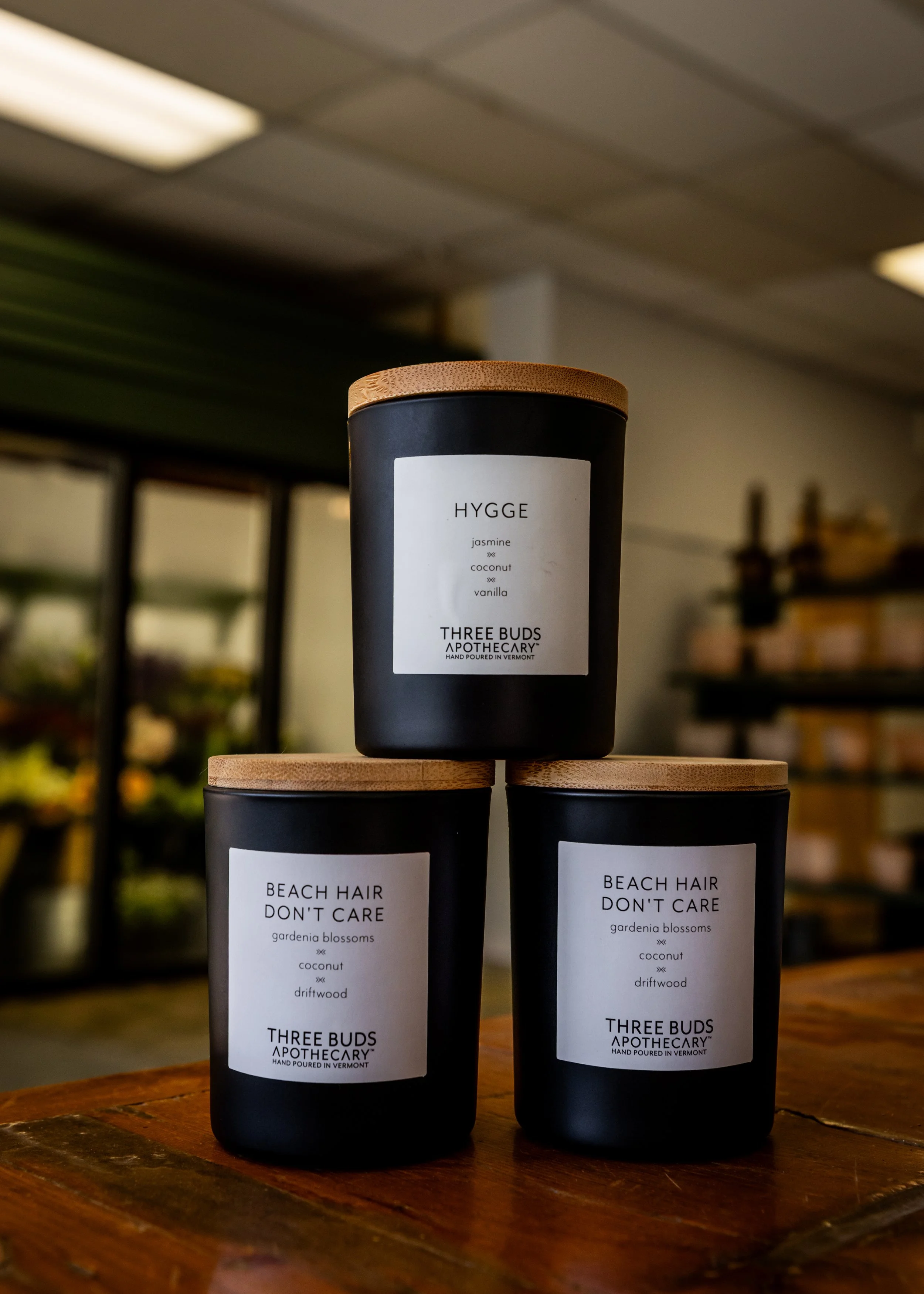 Stacked black candles with wooden lids on a wooden surface, labels reading 'HYGGE' with jasmine, coconut, vanilla, and 'BEACH HAIR DON'T CARE' with gardenia blossoms, coconut, driftwood, in a store with shelves and plants in the background.