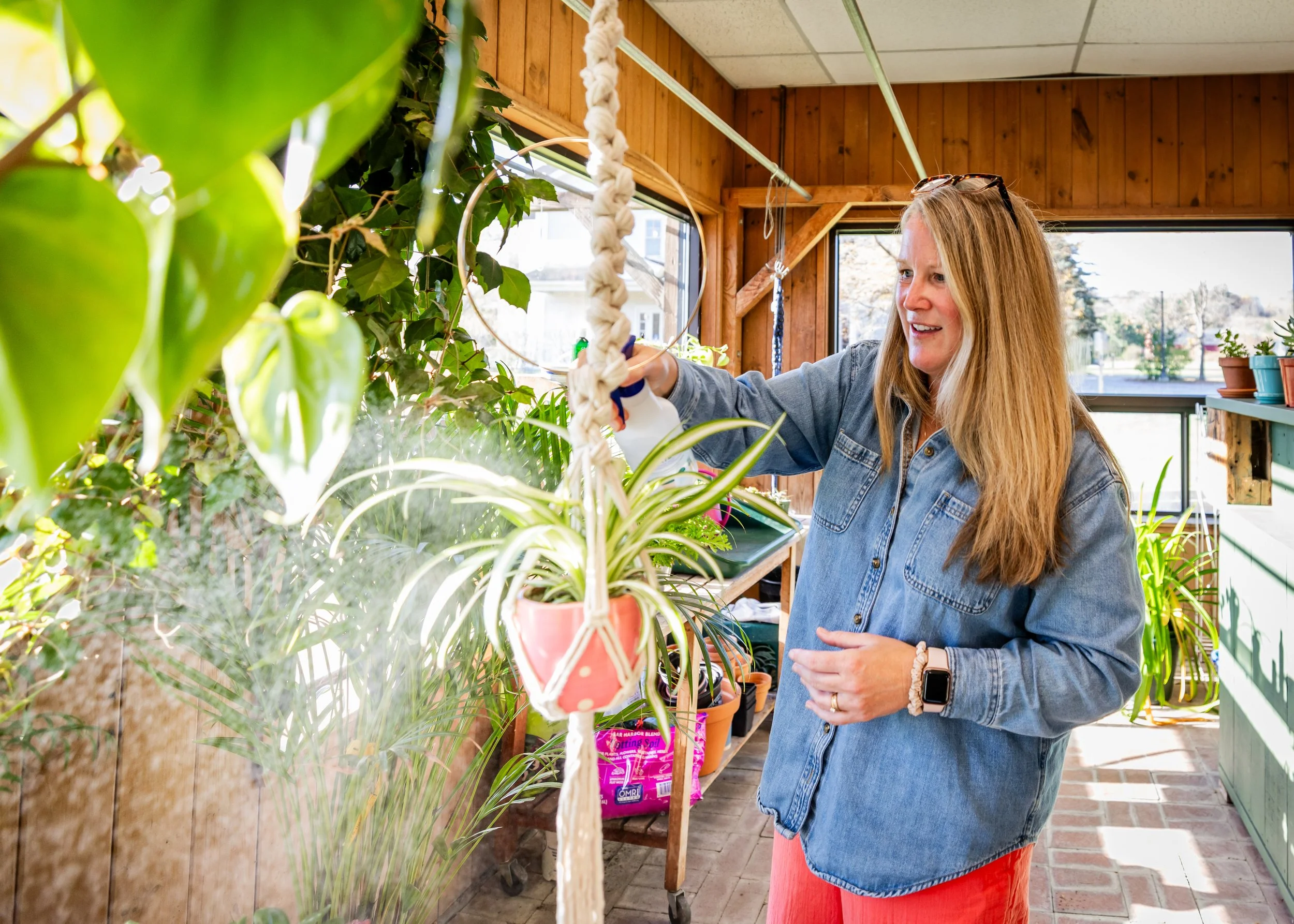 A woman watering plants with a spray bottle in a sunny indoor garden.