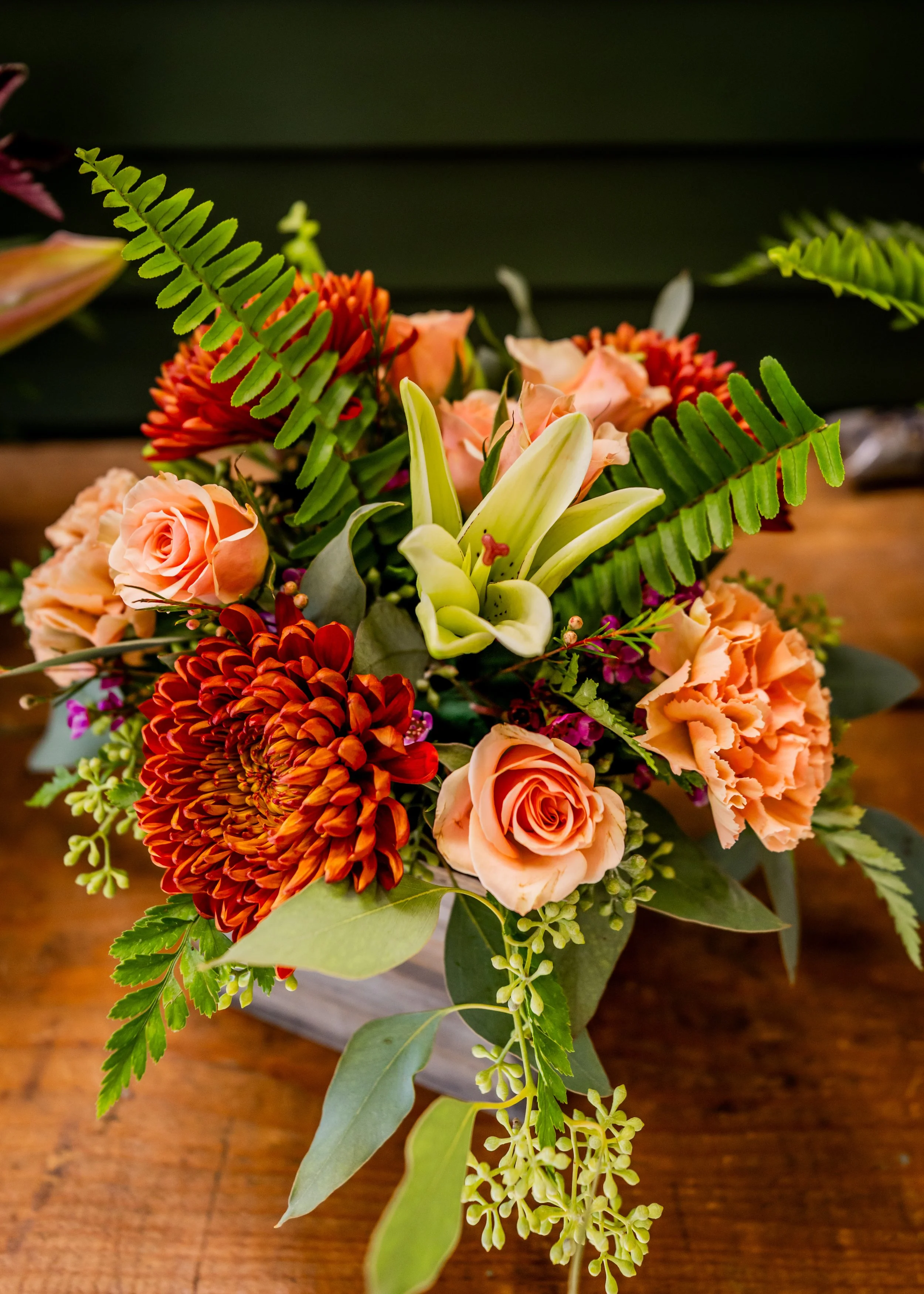 A colorful flower arrangement featuring peach roses, orange chrysants, white lilies, and green fern leaves, placed on a wooden surface.
