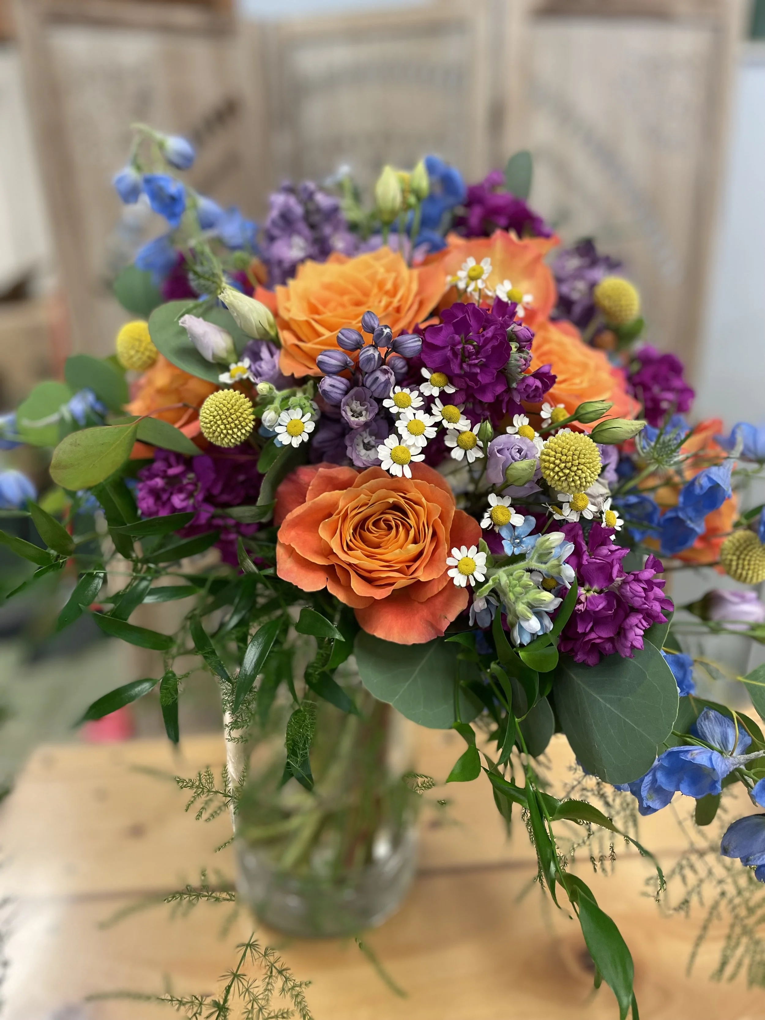 Colorful bouquet of orange roses, purple snapdragons, blue delphiniums, white daisies, and other mixed flowers in a glass vase on a wooden table.