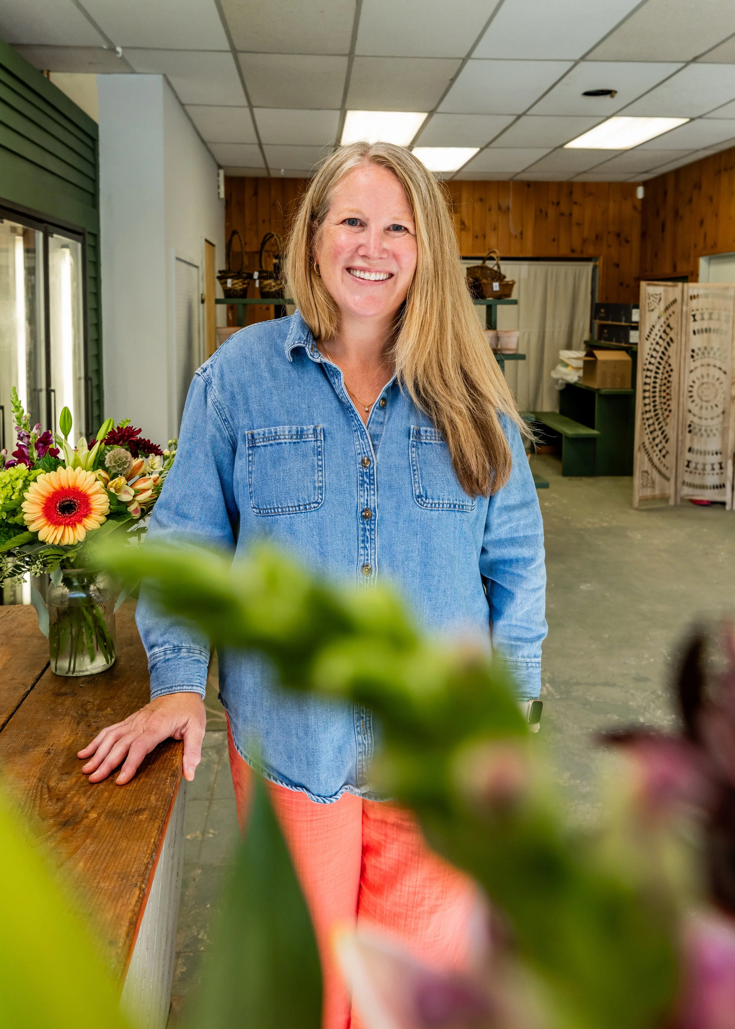 A smiling woman with long blonde hair wearing a denim shirt and red pants, standing behind a wooden counter with a colorful flower bouquet in a store or workshop setting.