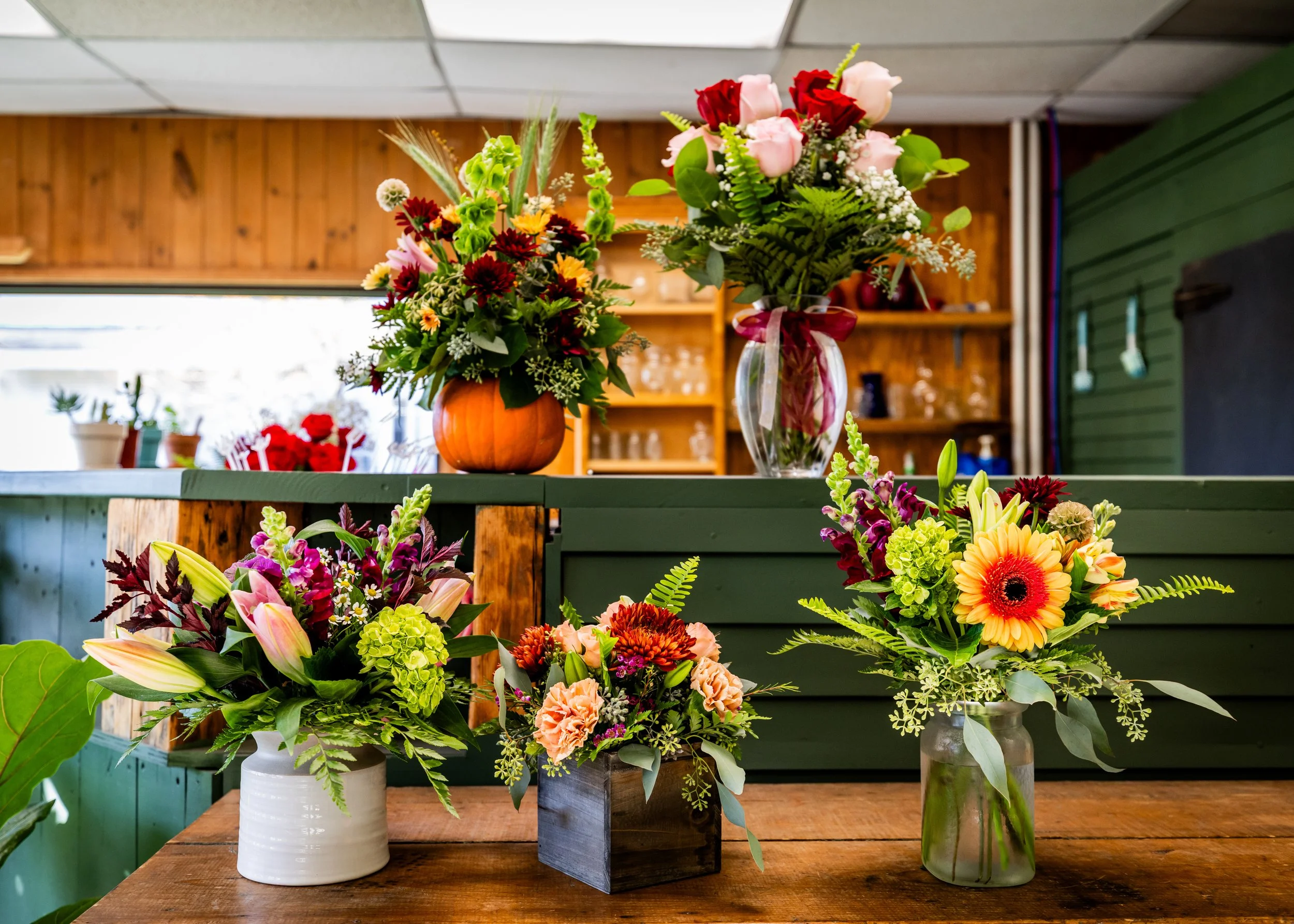 Multiple colorful flower arrangements displayed on wooden and green surfaces, with a background of a wooden wall and shelf.