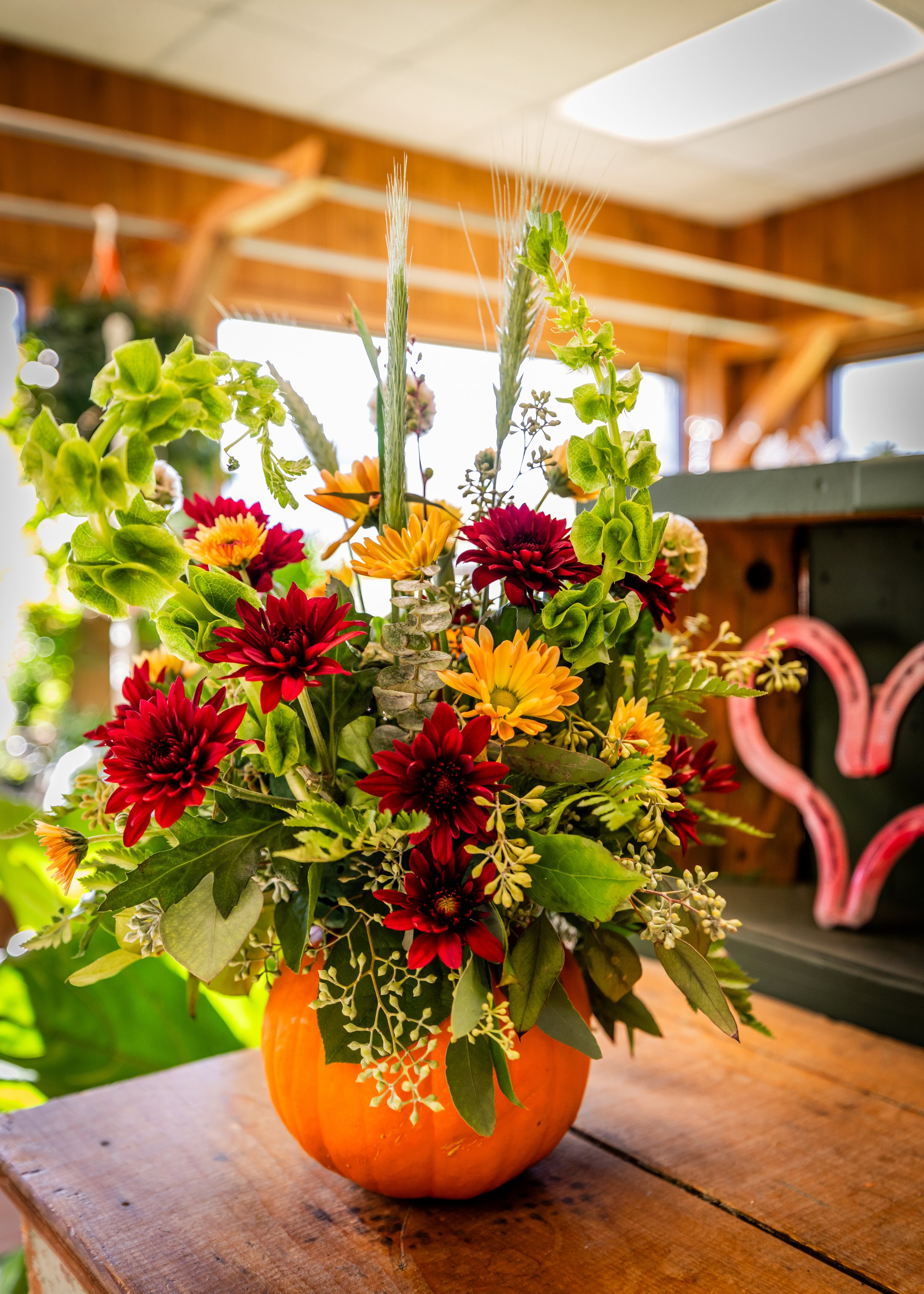 A fall-themed floral arrangement in a pumpkin-shaped container, featuring red, yellow, and orange flowers with green leaves, placed on a wooden surface inside a room with wooden walls and large windows.