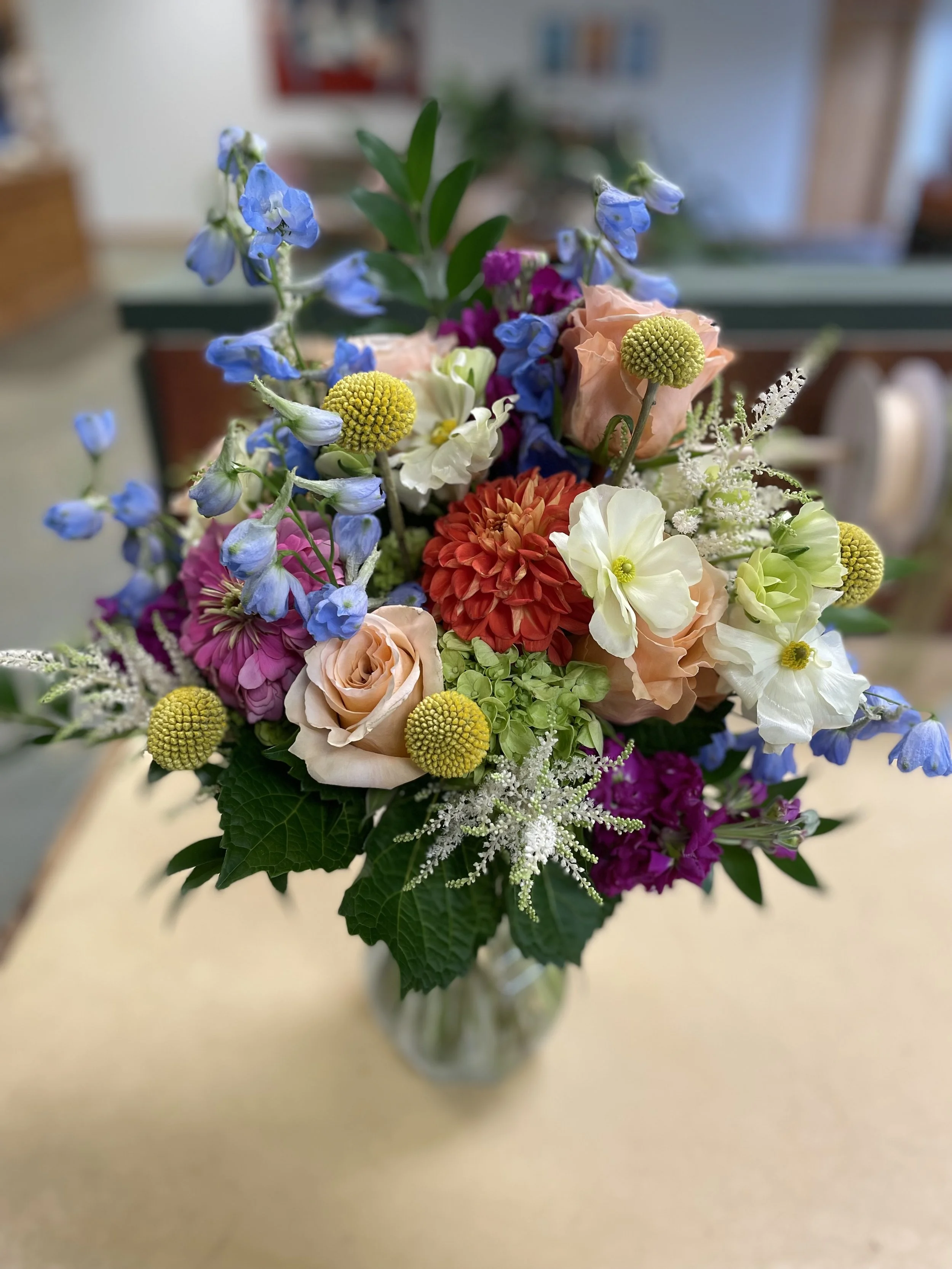 Colorful bouquet of various flowers including roses, dahlias, delphiniums, and other blossoms in a glass vase on a beige surface.