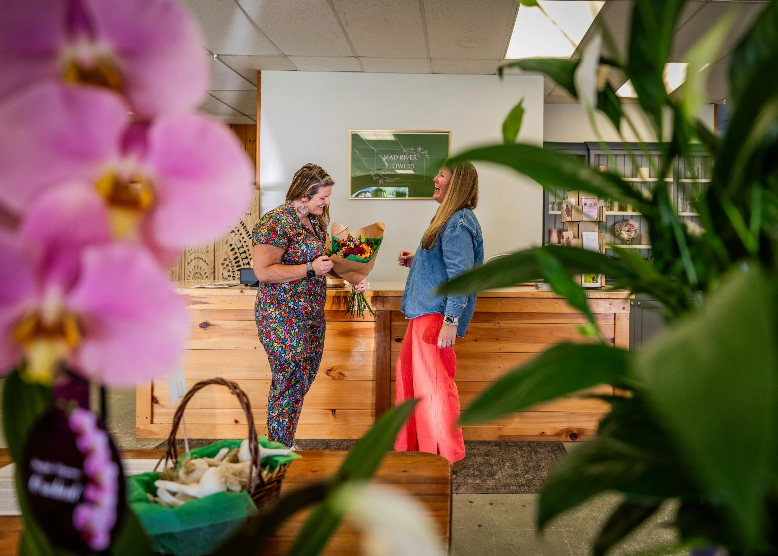Two women in a flower shop, one handing a bouquet of flowers to the other, seen through foregrounds of pink orchids and green leaves