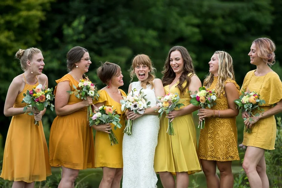 Bride in white dress surrounded by six bridesmaids in yellow dresses, all holding bouquets, outdoors with green trees in background.