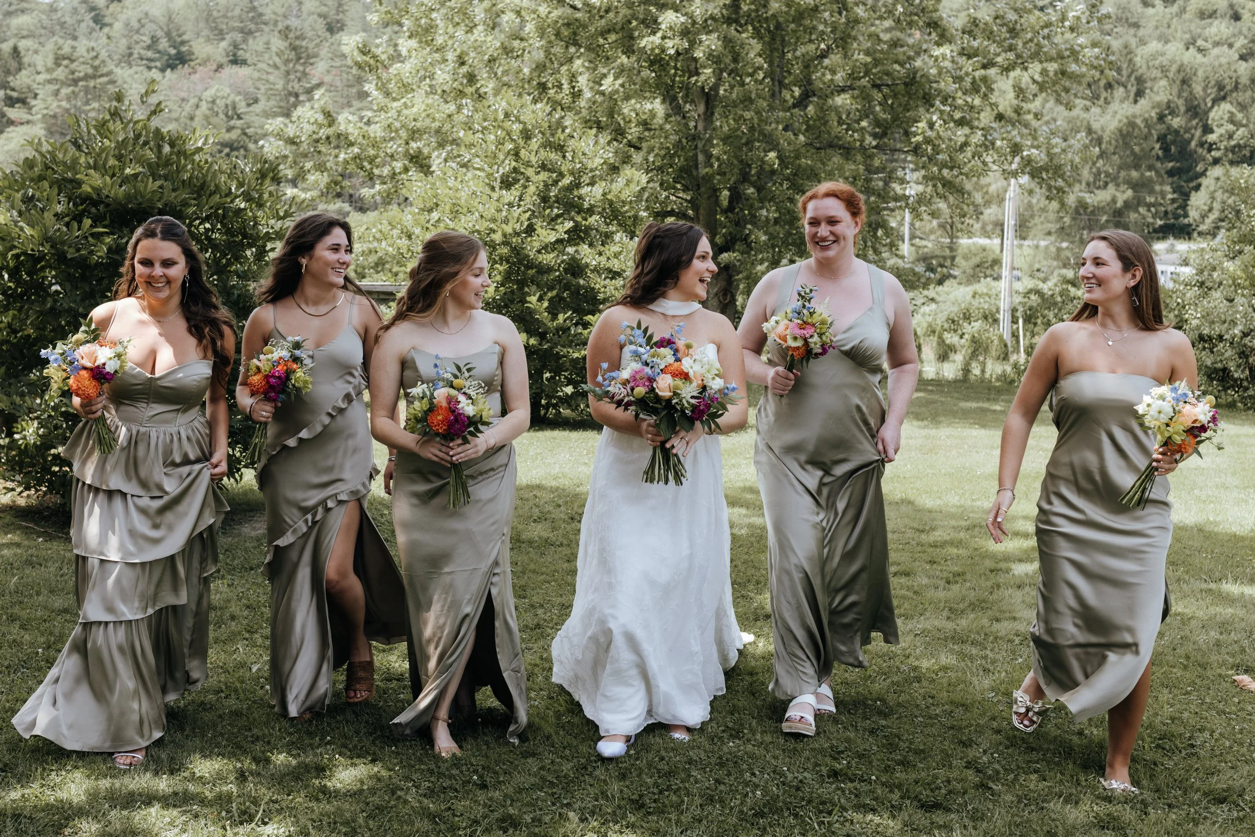 Seven women, including a bride in a white dress, walking outdoors on a grassy area with trees in the background, holding colorful bouquets and smiling.
