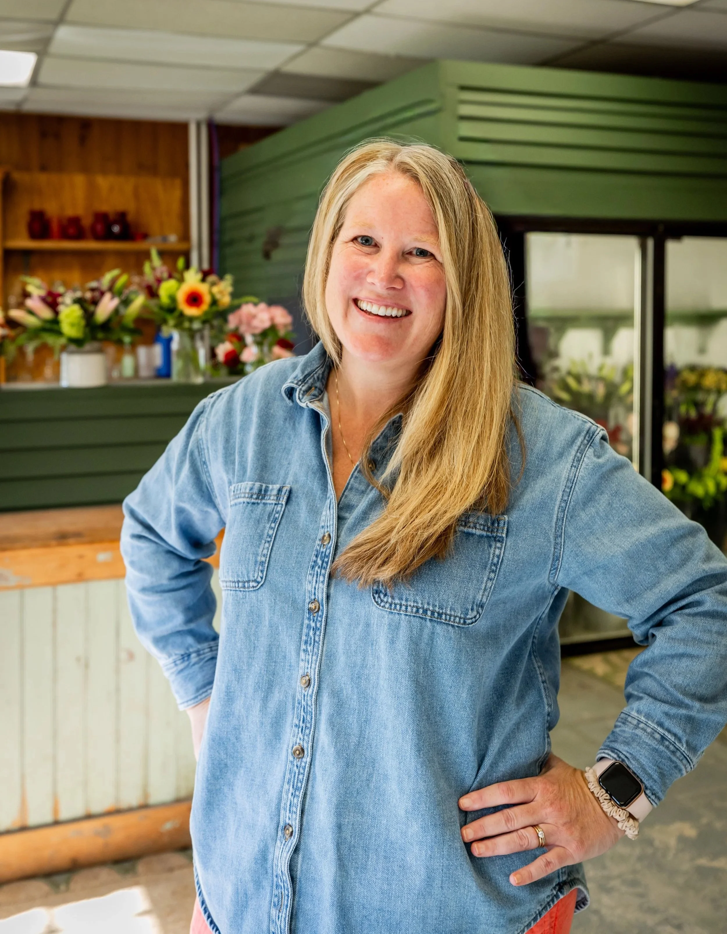 A smiling woman with long blond hair wearing a denim shirt and smartwatch, standing inside a flower shop.