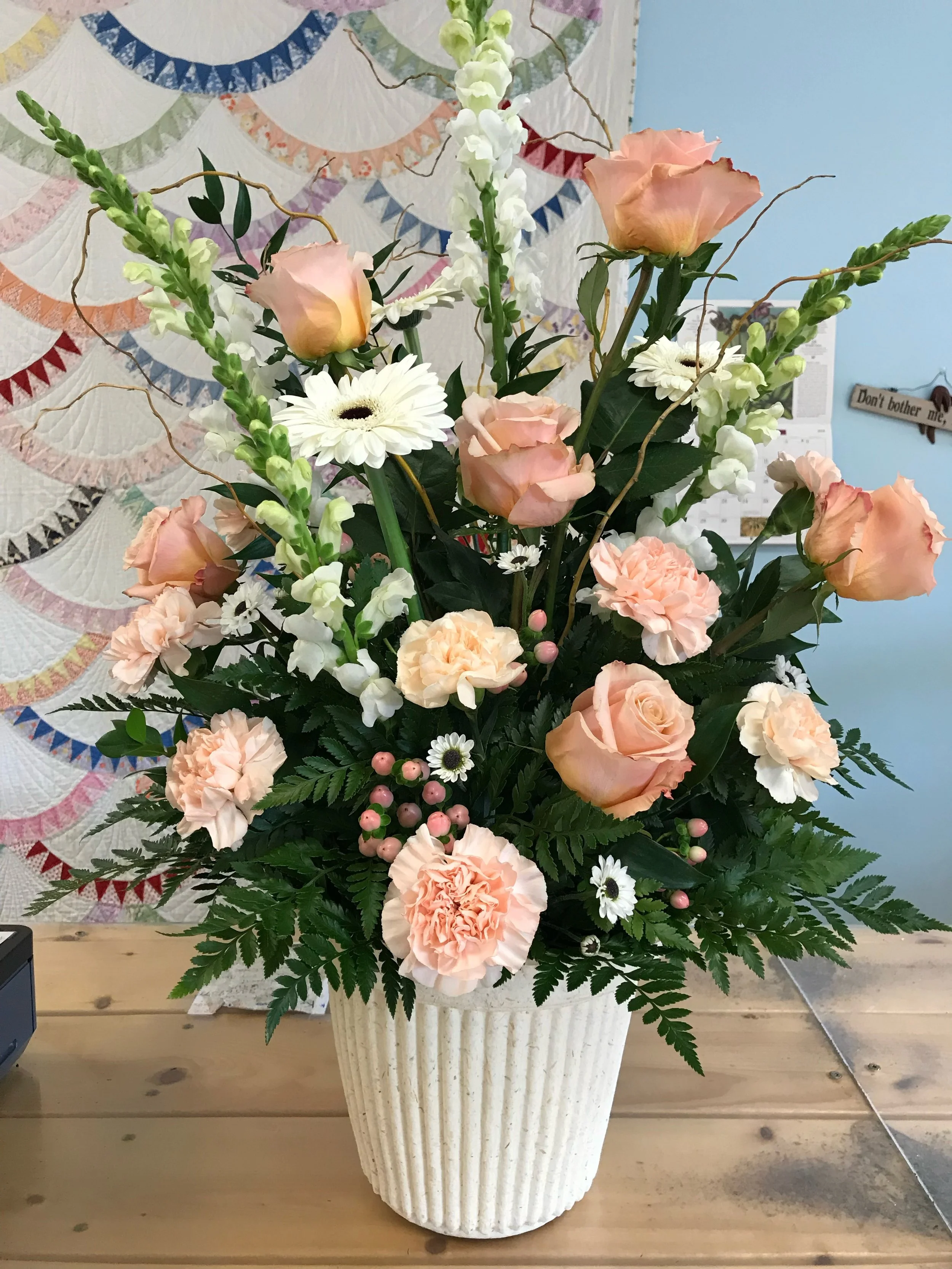 A floral arrangement with pink roses, white gerbera daisies, peach carnations, white snapdragons, small pink berries, and green ferns in a white ribbed ceramic vase on a wooden table.