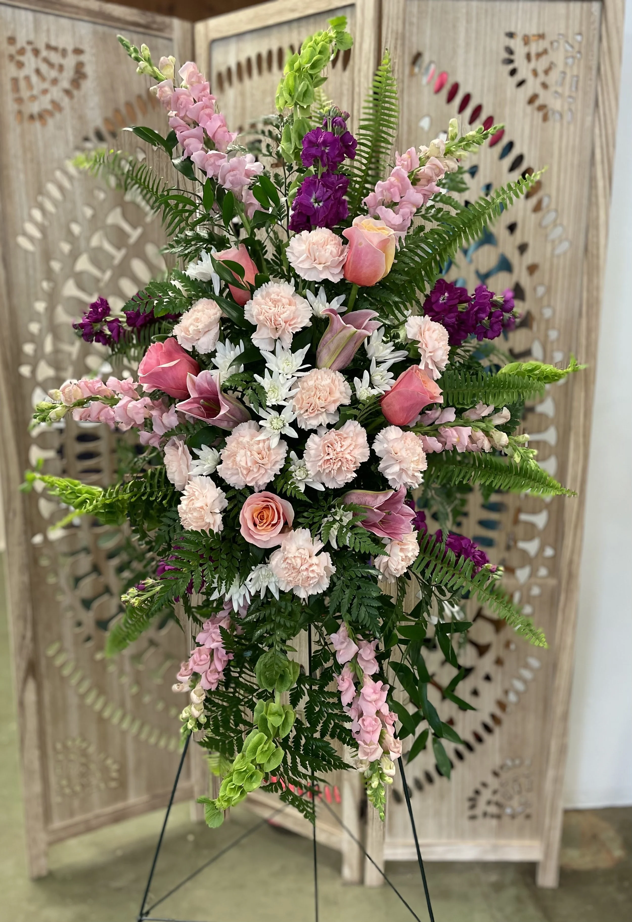 A colorful flower arrangement with pink, purple, white, and peach flowers, surrounded by green fern leaves, set on a stand in front of a decorative wooden screen.