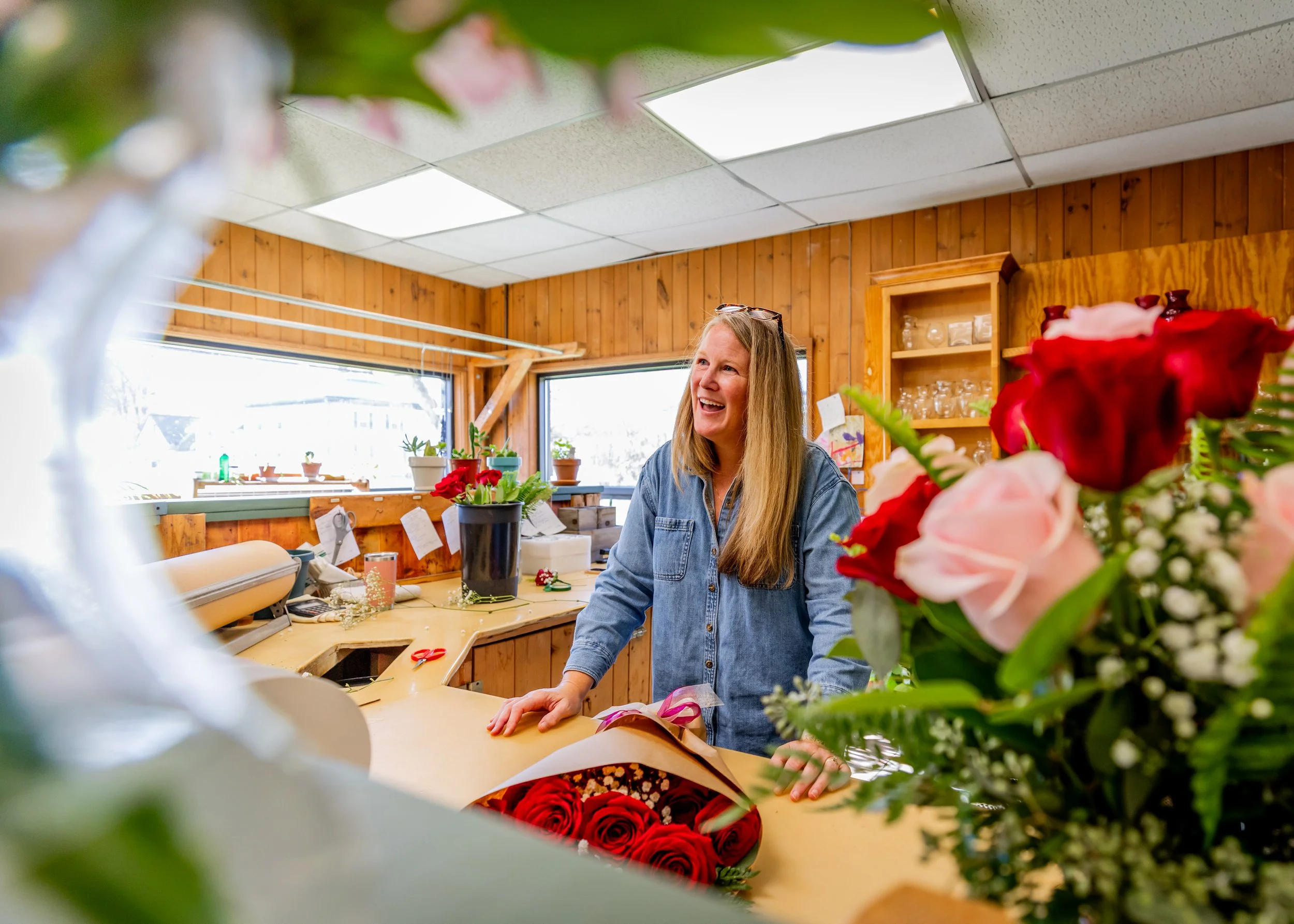 A woman with long hair, wearing a denim shirt, smiling and standing behind a counter with colorful flower arrangements, inside a wood-paneled room with large windows.