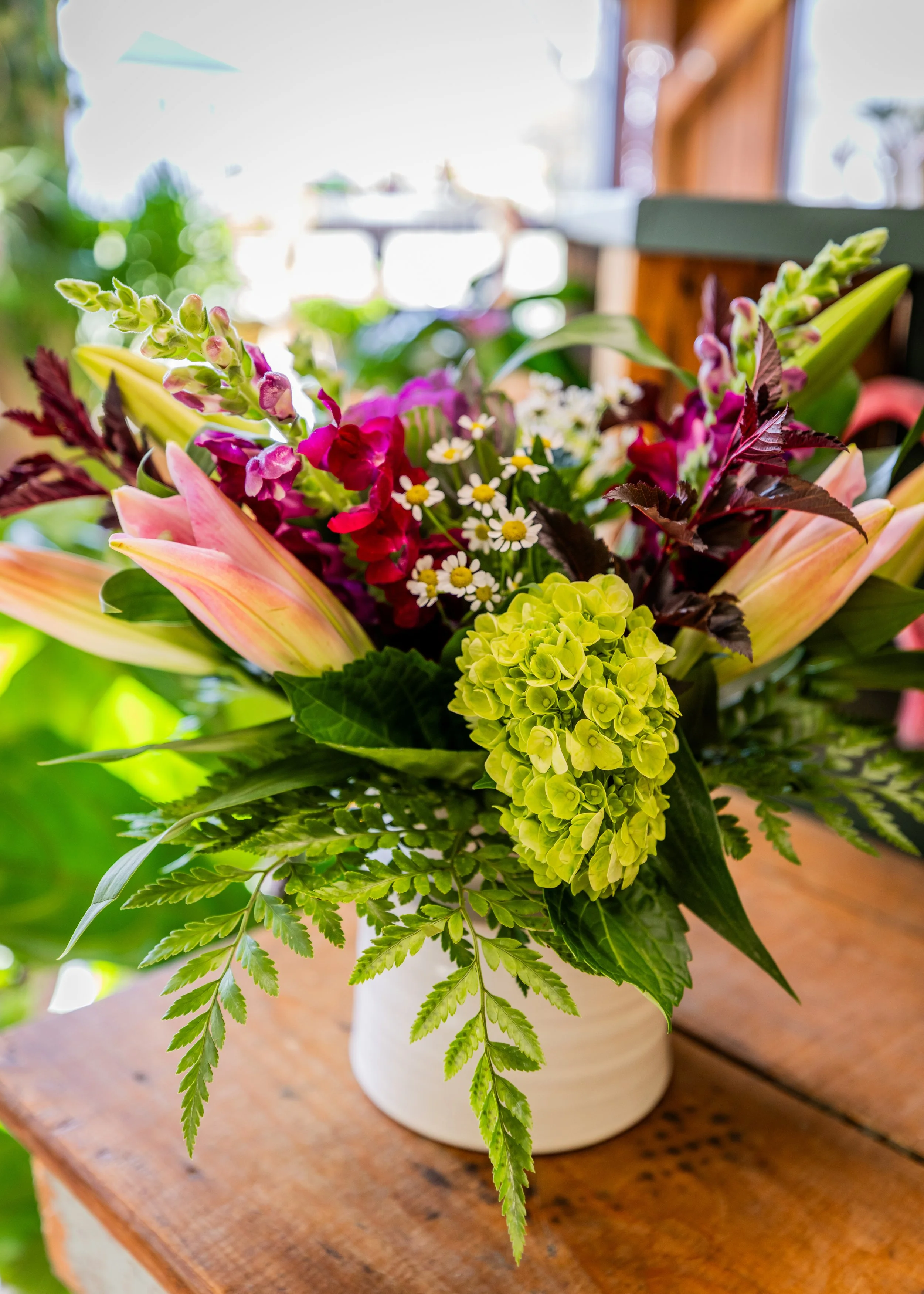 Colorful mixed flower bouquet in a white vase on wooden surface, with a blurred outdoor garden background.