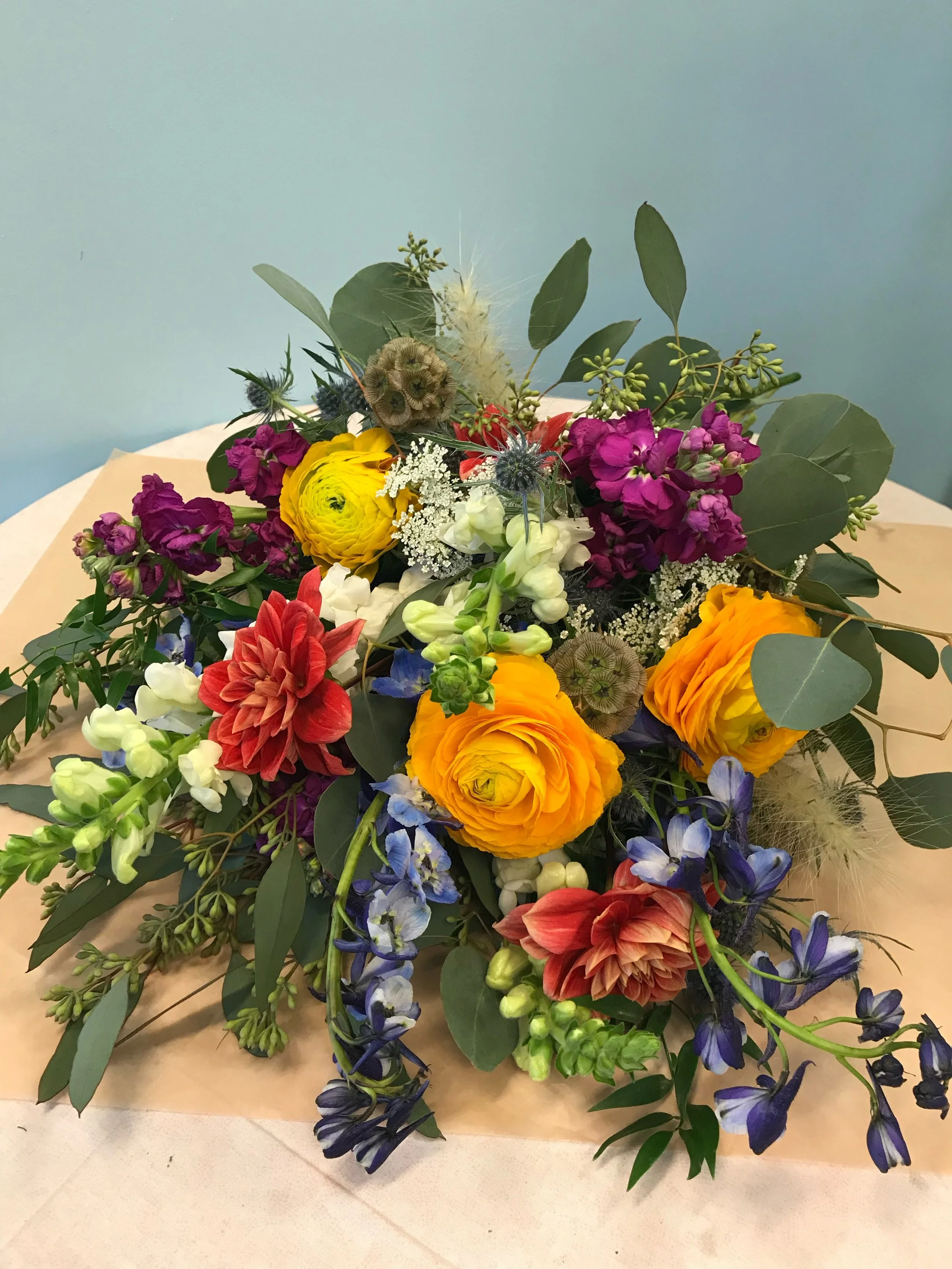 A colorful bouquet of fresh flowers including yellow ranunculus, red dahlias, purple stock, white snapdragons, blue delphiniums, and greenery on a beige paper background.