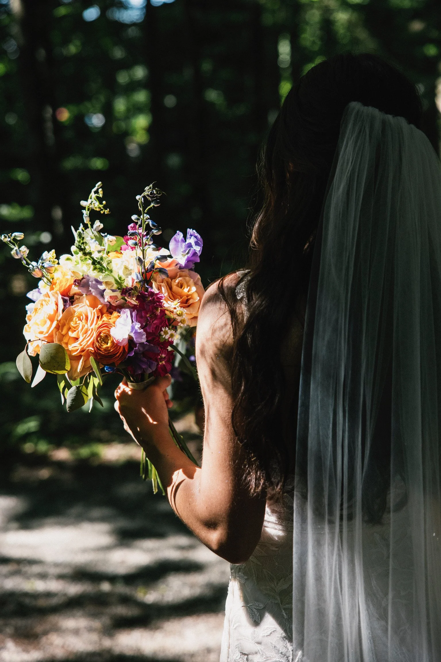 Bride holding a colorful bouquet of flowers, with her face partially obscured by a veil, in a wooded outdoor setting.