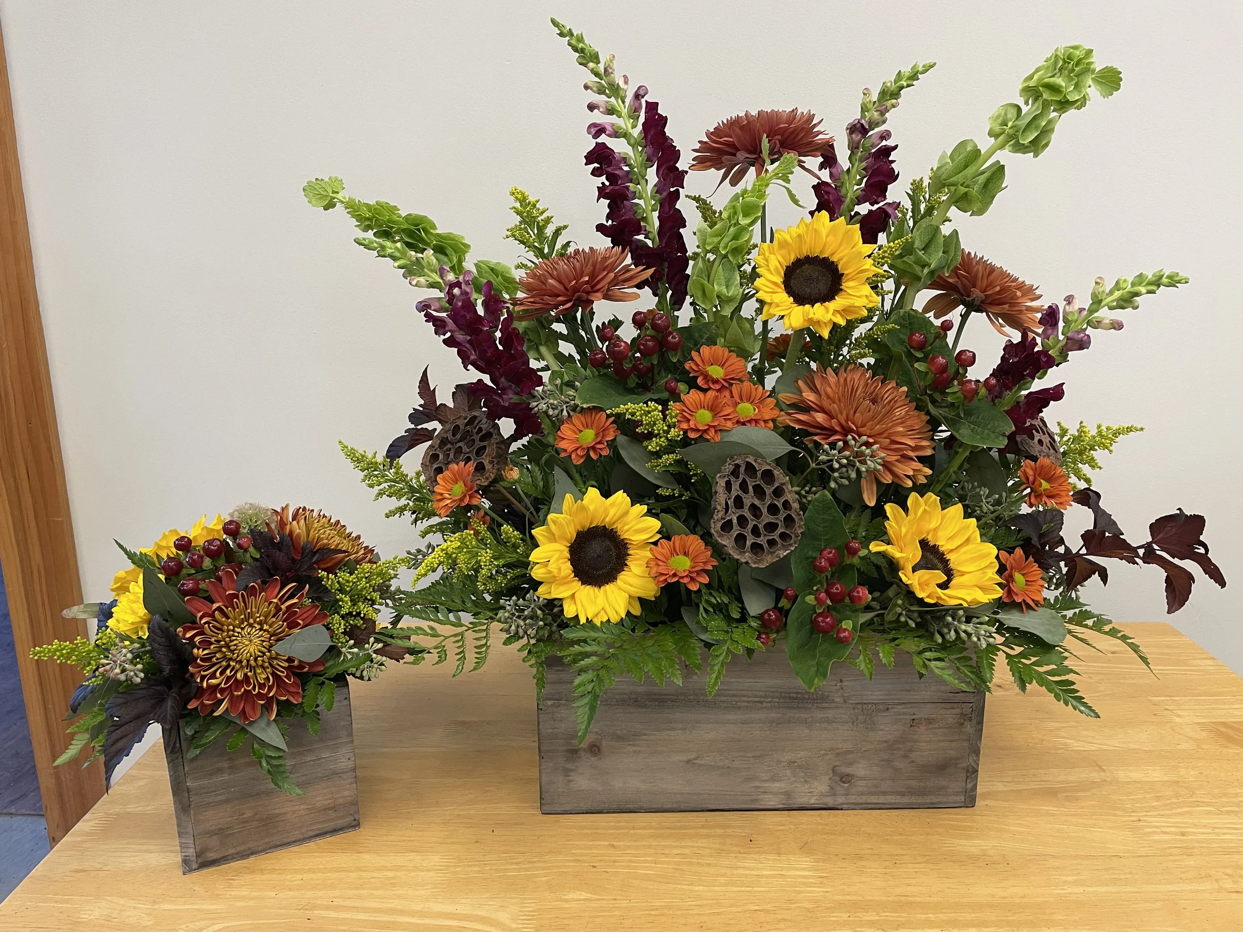 Two wooden planters filled with colorful fall flowers and foliage, including sunflowers, orange chrysanthemums, purple gladiolus, and burgundy berries, placed on a wooden table against a plain background.