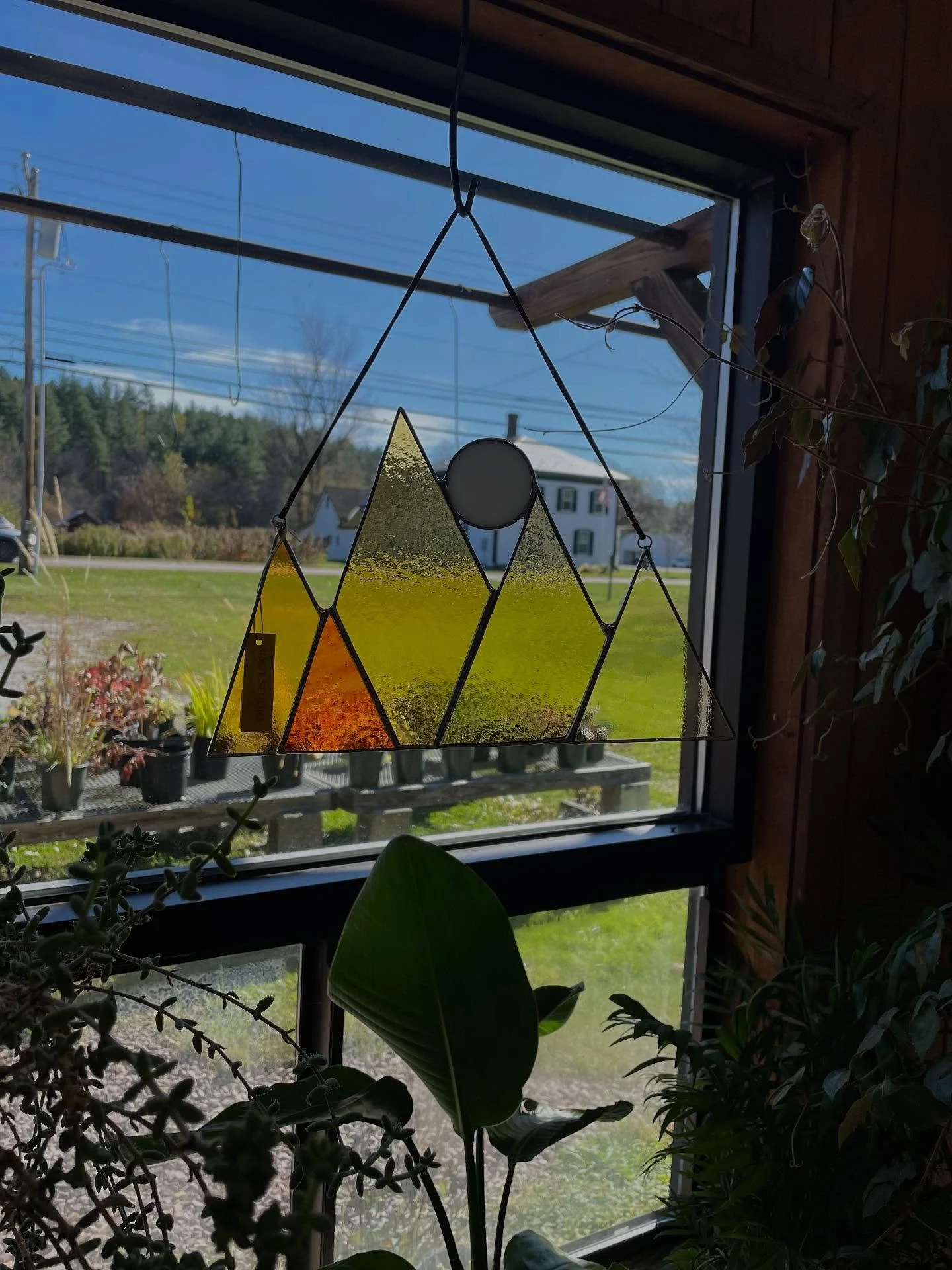 A stained glass window hangs in front of a large window. The stained glass design is of a mountain landscape with yellow, orange, and green colors, and a white circle representing the sun or moon.