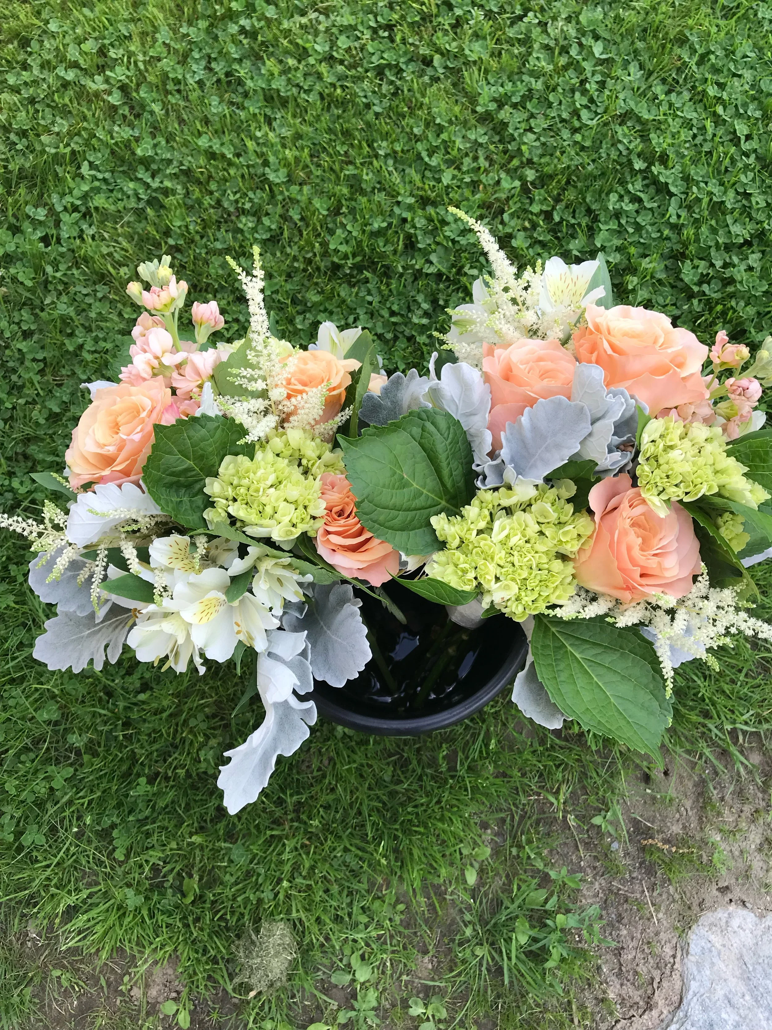 A bouquet of peach roses, white alstroemeria, green hydrangeas, and assorted greenery in a black vase placed on grass.