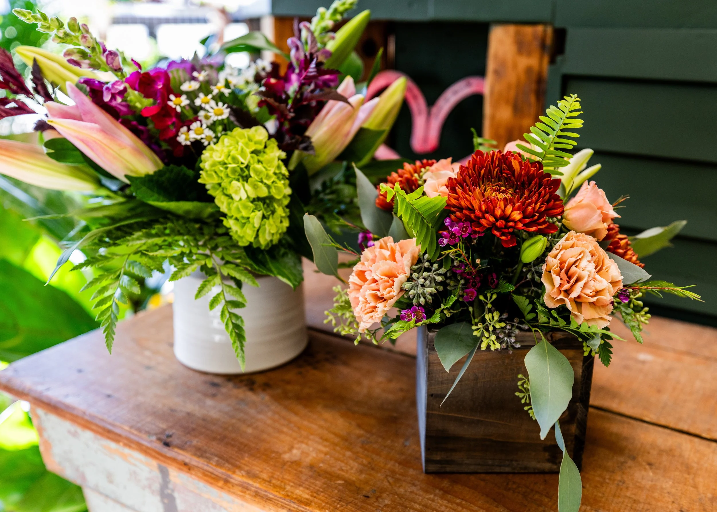 Two different flower arrangements on a wooden surface, one in a white ceramic pot with lilies and greenery, and the other in a wooden box with peach, red, and purple flowers and green foliage.