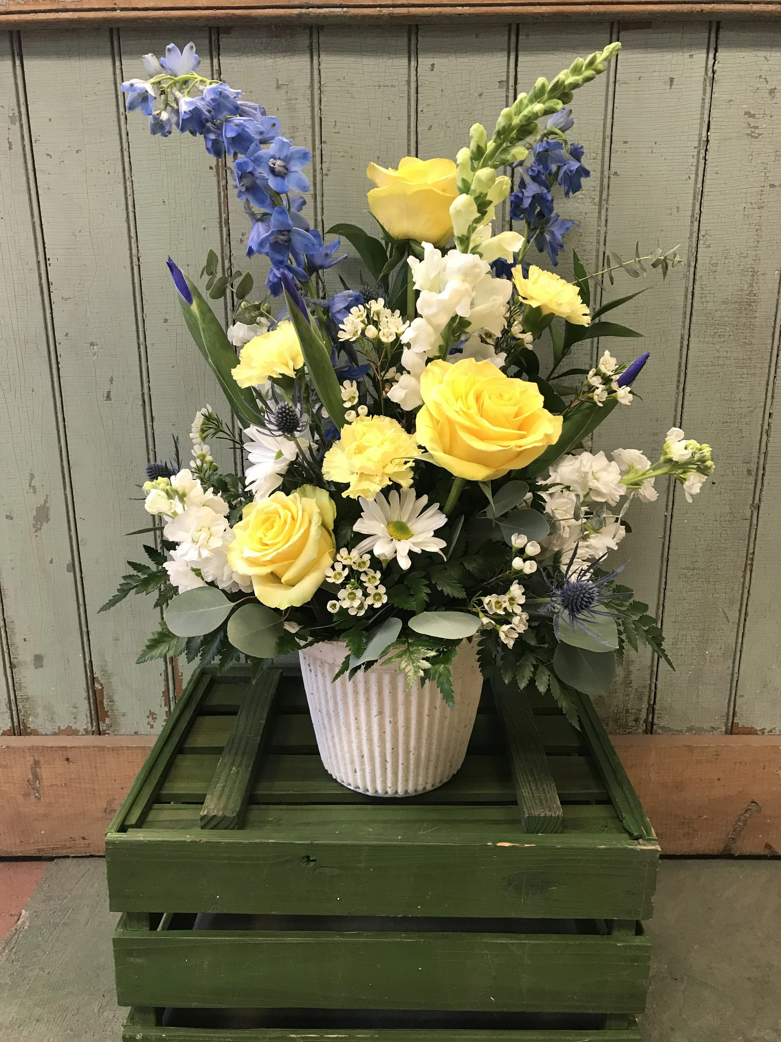 A bouquet of yellow roses, white daisies, blue delphiniums, and other flowers in a ribbed white ceramic pot, placed on a green wooden crate against a wooden wall background.