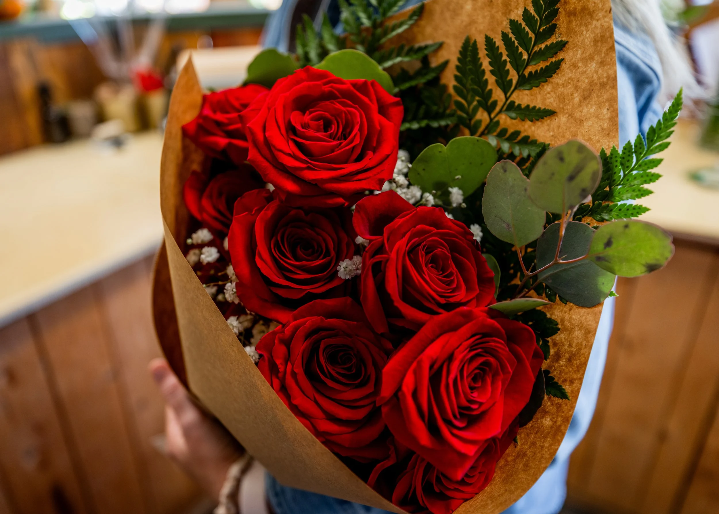 A bouquet of red roses with green leaves and white baby's breath wrapped in brown paper being held by a person.