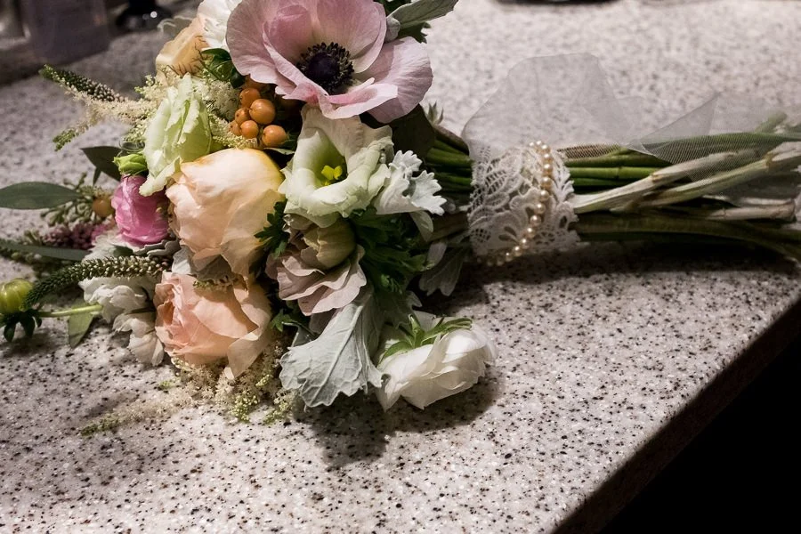 A bouquet of assorted flowers, including pink, white, and peach roses, is resting on a speckled gray countertop, tied with a lace ribbon and pearl embellishments.