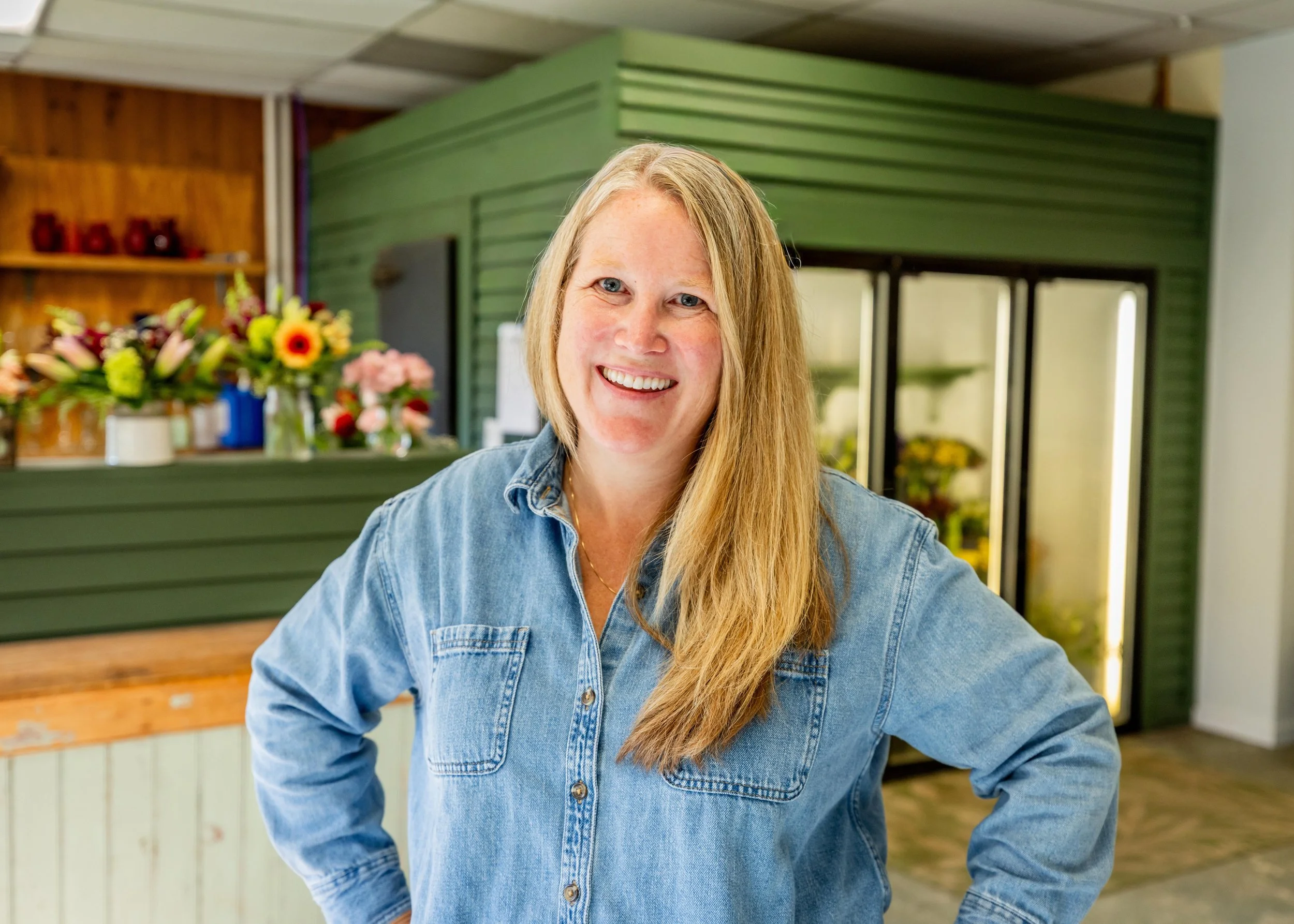 Smiling woman with long blonde hair wearing a blue denim shirt standing in a flower shop.