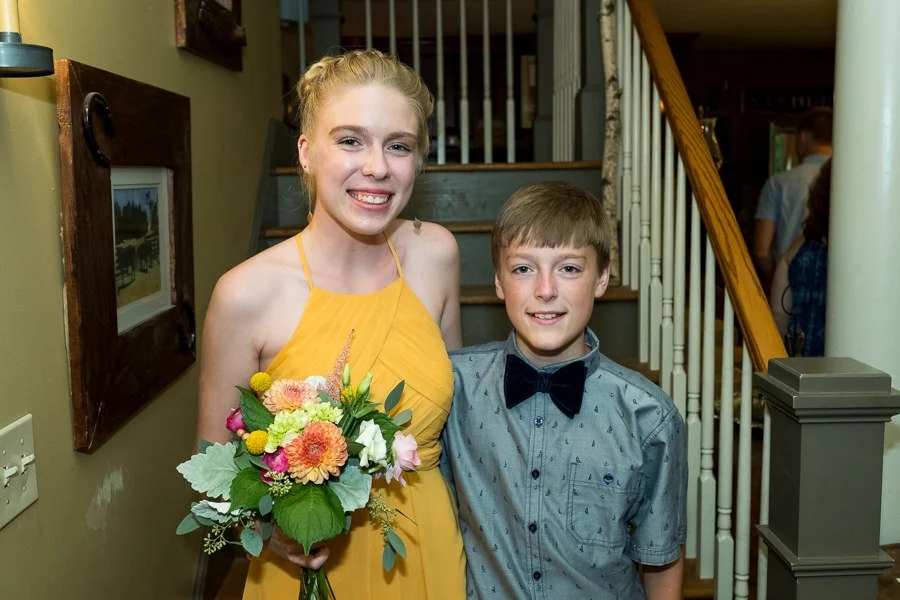 A young woman in a yellow dress holding a bouquet of flowers standing next to a young boy in a gray shirt with a black bow tie, smiling in a home interior with stairs in the background.