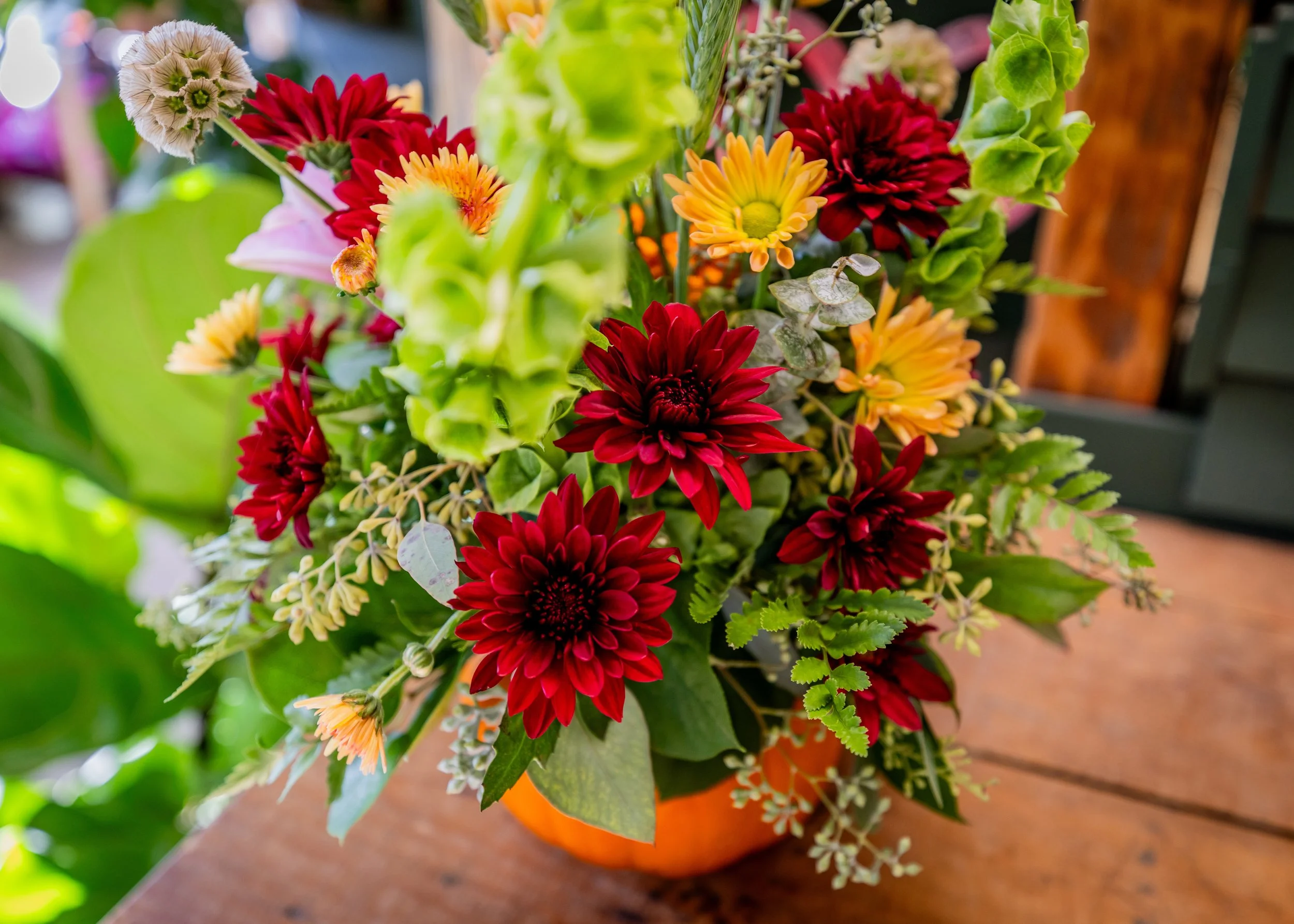A colorful bouquet of various flowers, including red, orange, and peach blossoms, arranged in an orange pot on a wooden surface with blurred greenery in the background.