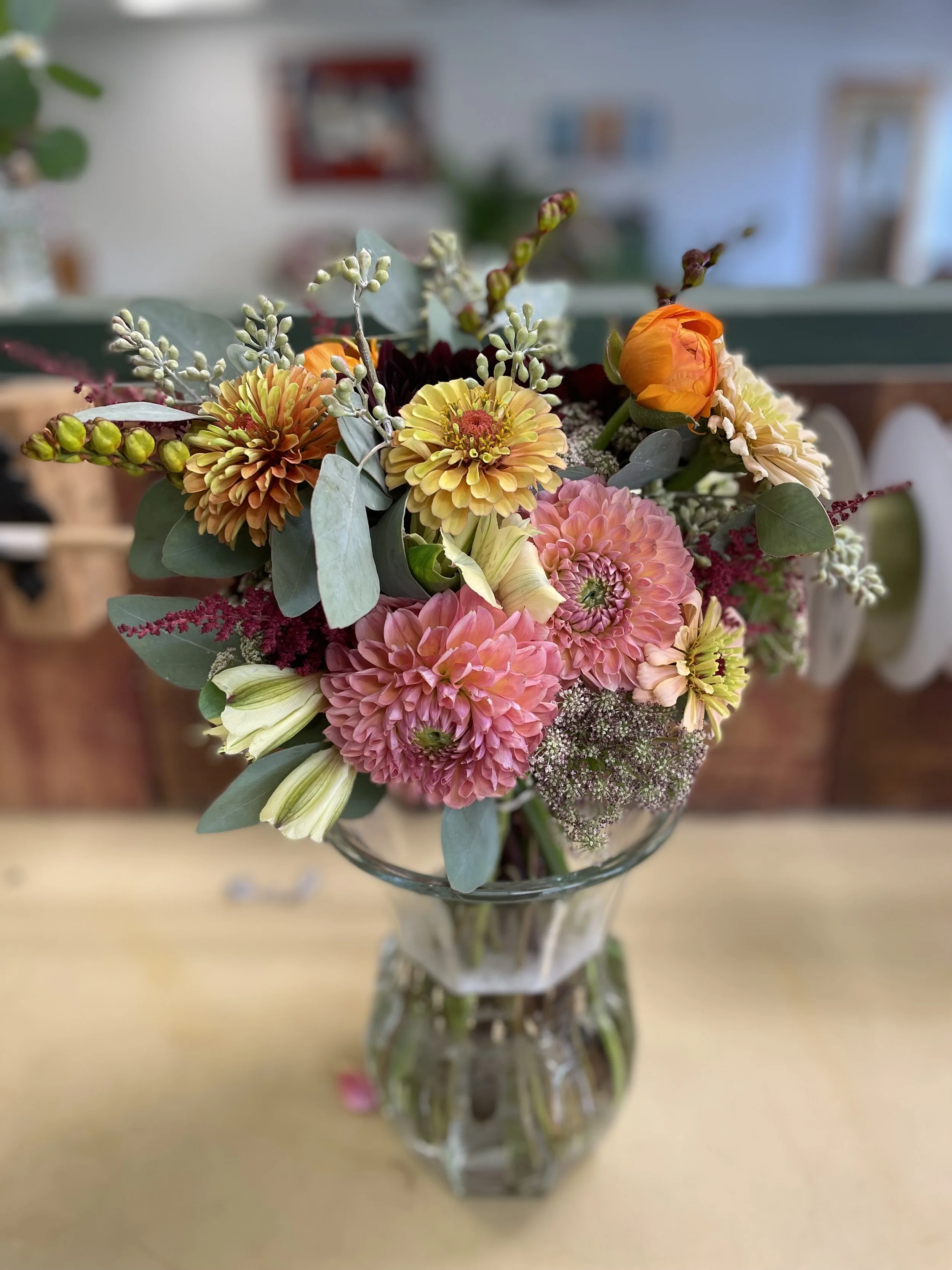 A colorful flower bouquet in a glass vase on a table, featuring pink, yellow, orange, and white flowers with green leaves and foliage.