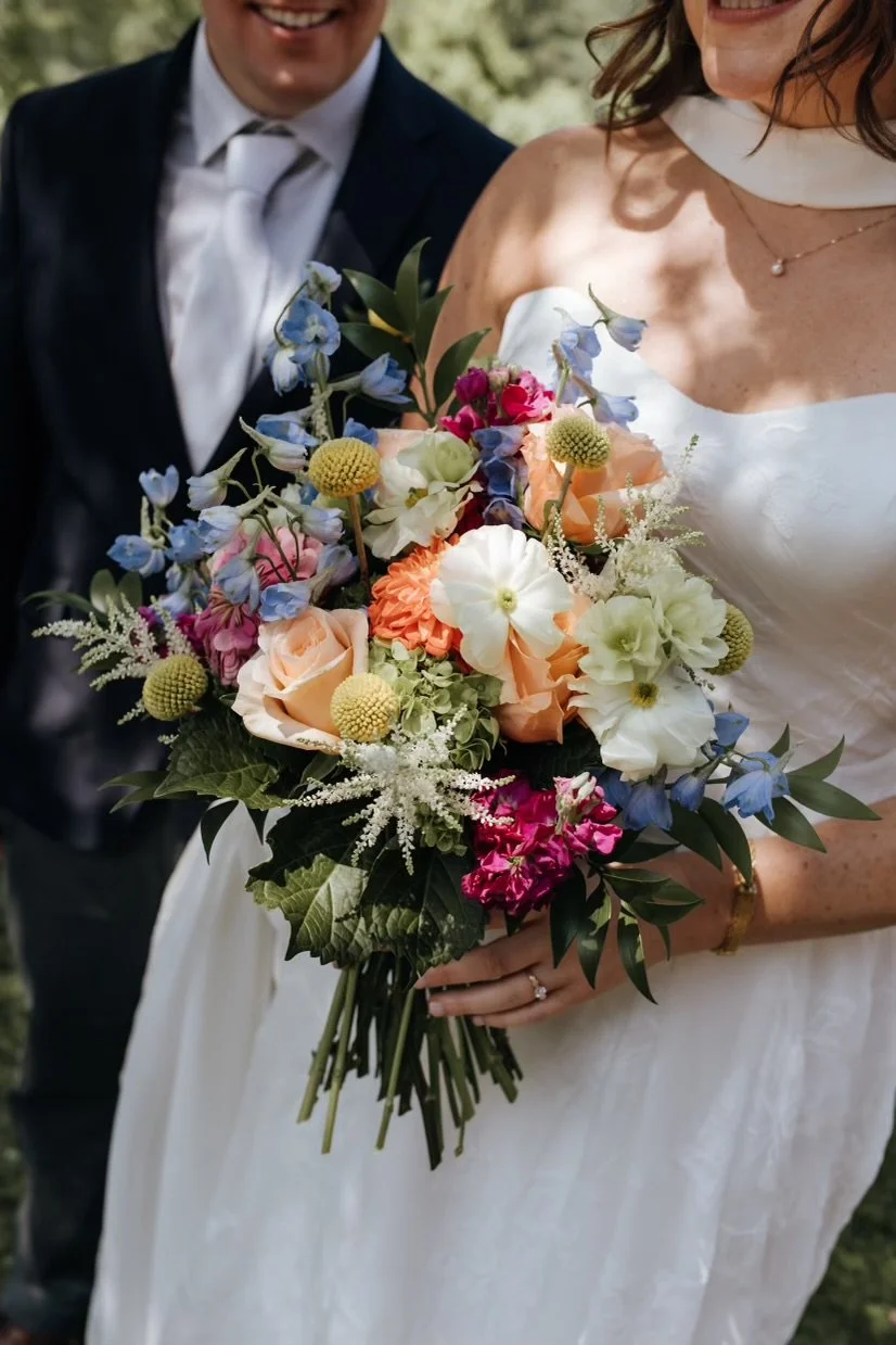 Close-up view of a bride holding a colorful bouquet of flowers, with a groom in a dark suit and white shirt slightly in background.