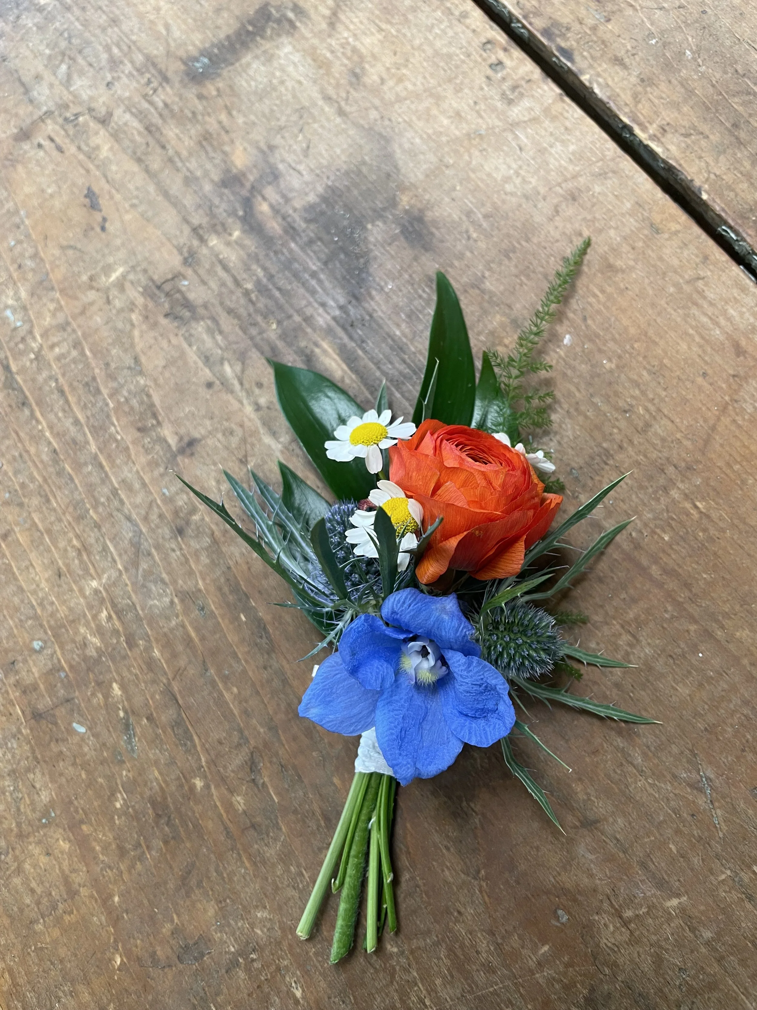 A small bouquet of mixed flowers including orange roses, blue iris, small white daisies, and greenery, placed on a wooden surface.