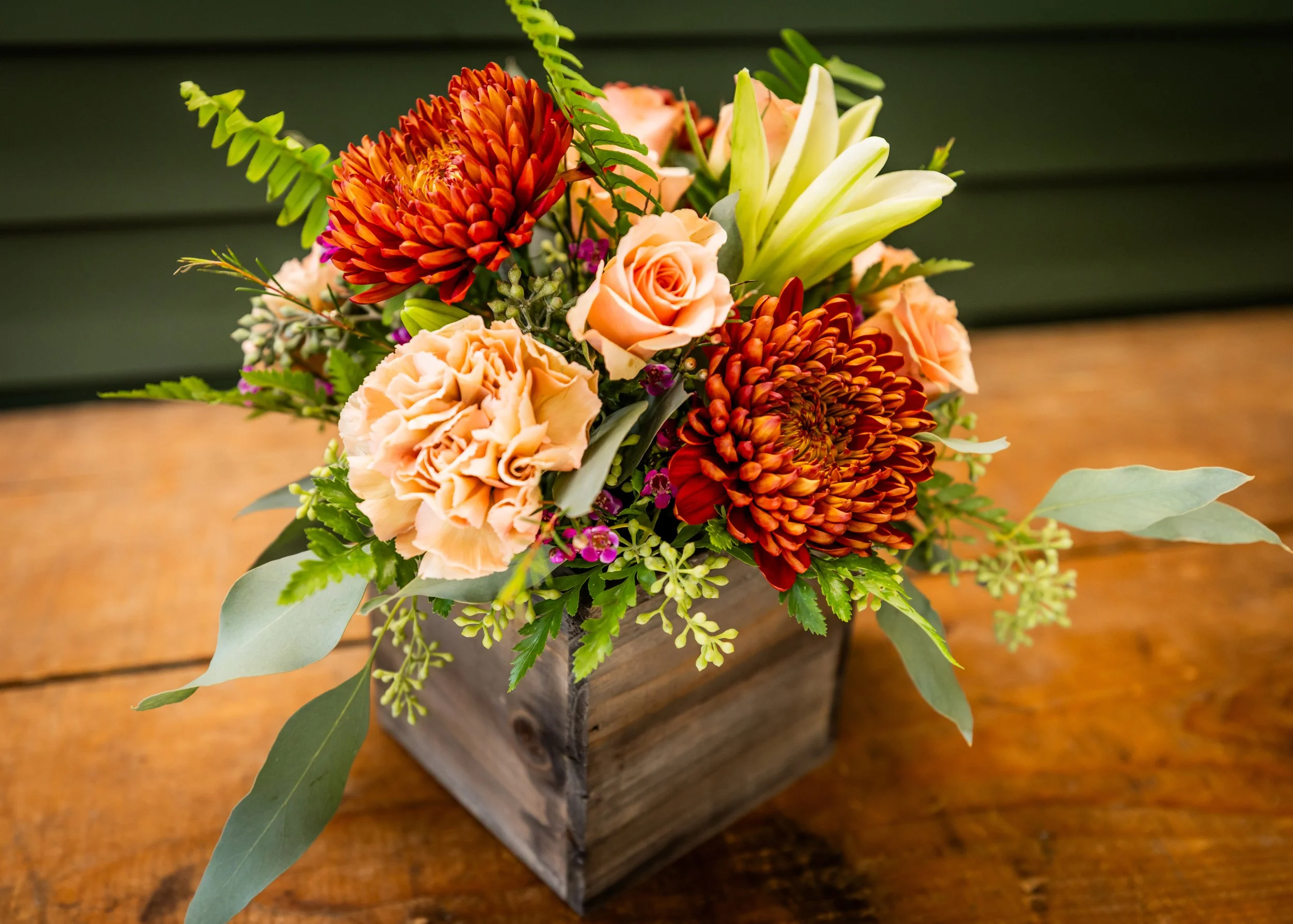A floral arrangement in a wooden square container with peach roses, orange dahlias, white lilies, green leaves, and small purple flowers on a wooden table.