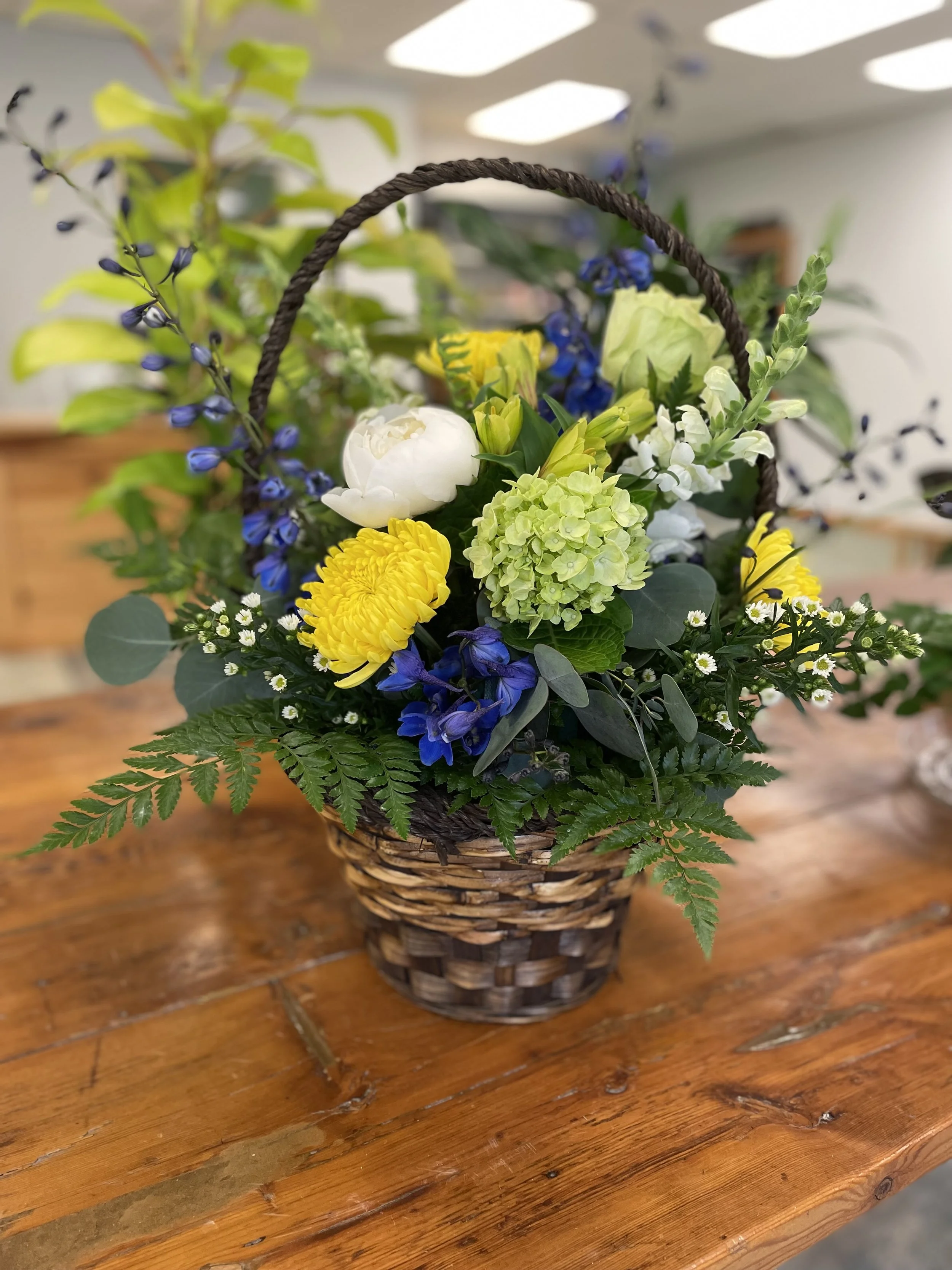 A wicker basket filled with various colorful flowers, including white, yellow, green, and blue blooms, sits on a wooden table.