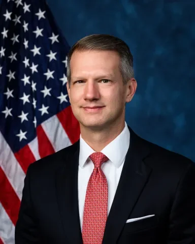 A man in a suit and red tie standing in front of an American flag.