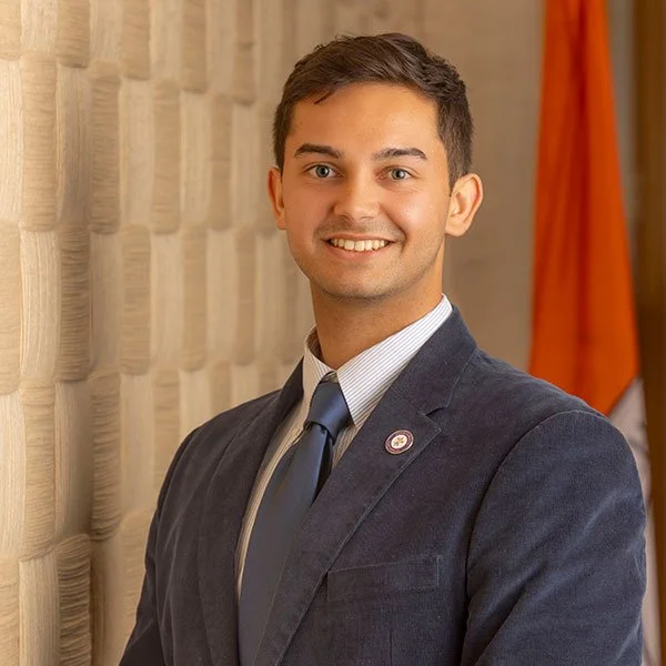 Young man in a suit smiling, standing indoors with a textured wall and a flag in the background.