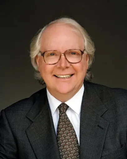 A smiling elderly man with glasses, wearing a dark suit, white shirt, and patterned tie, against a dark background.