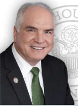 A middle-aged man with a bald head, wearing a dark suit, white shirt, and green tie, smiling at the camera. The background includes a blurred emblem or crest.
