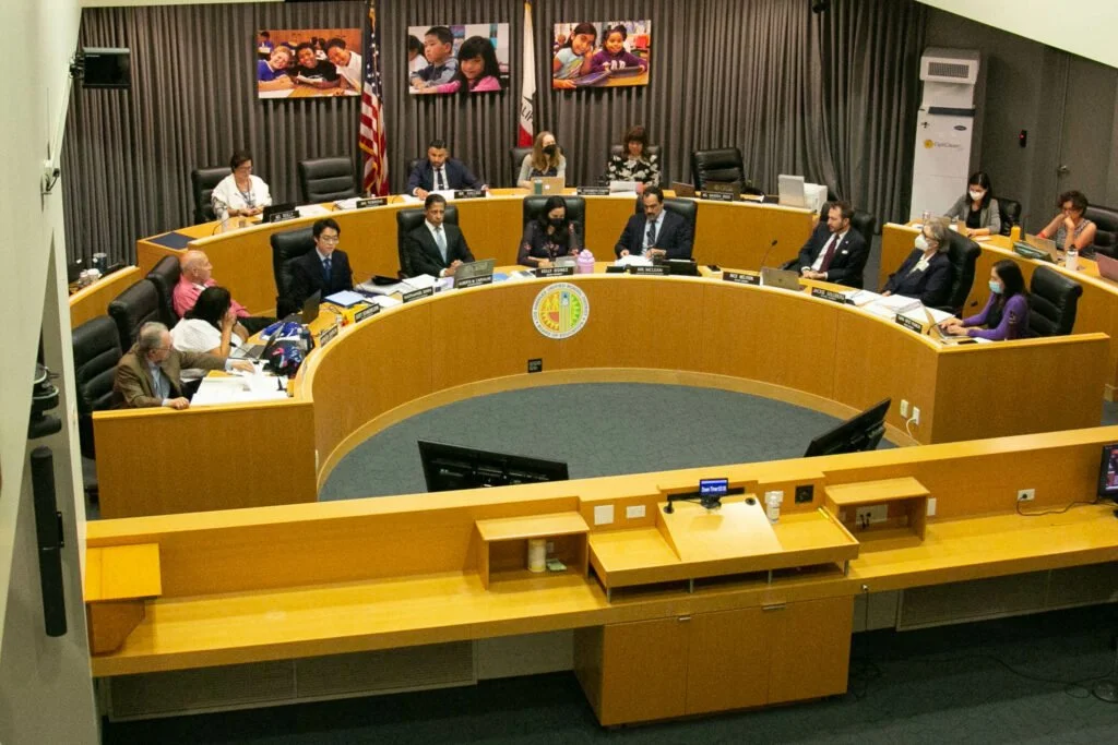 A group of people in a formal meeting or hearing room with curved desk, some wearing face masks, and large photos of children on the dark curtain backdrop.