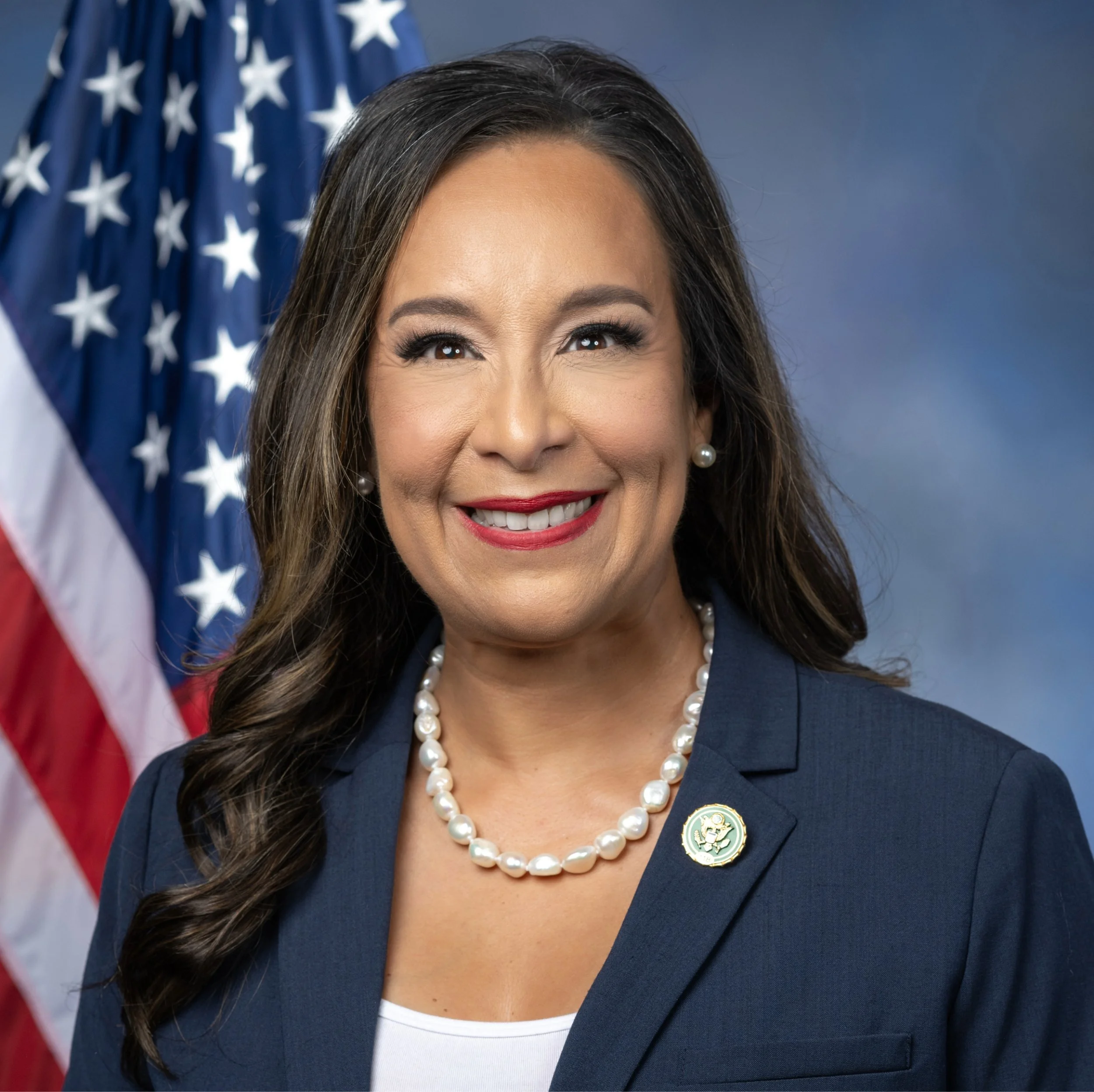 Partial portrait of a woman with long brown hair, wearing a pearl necklace, a navy blazer with a badge, and pearl earrings, standing in front of an American flag.