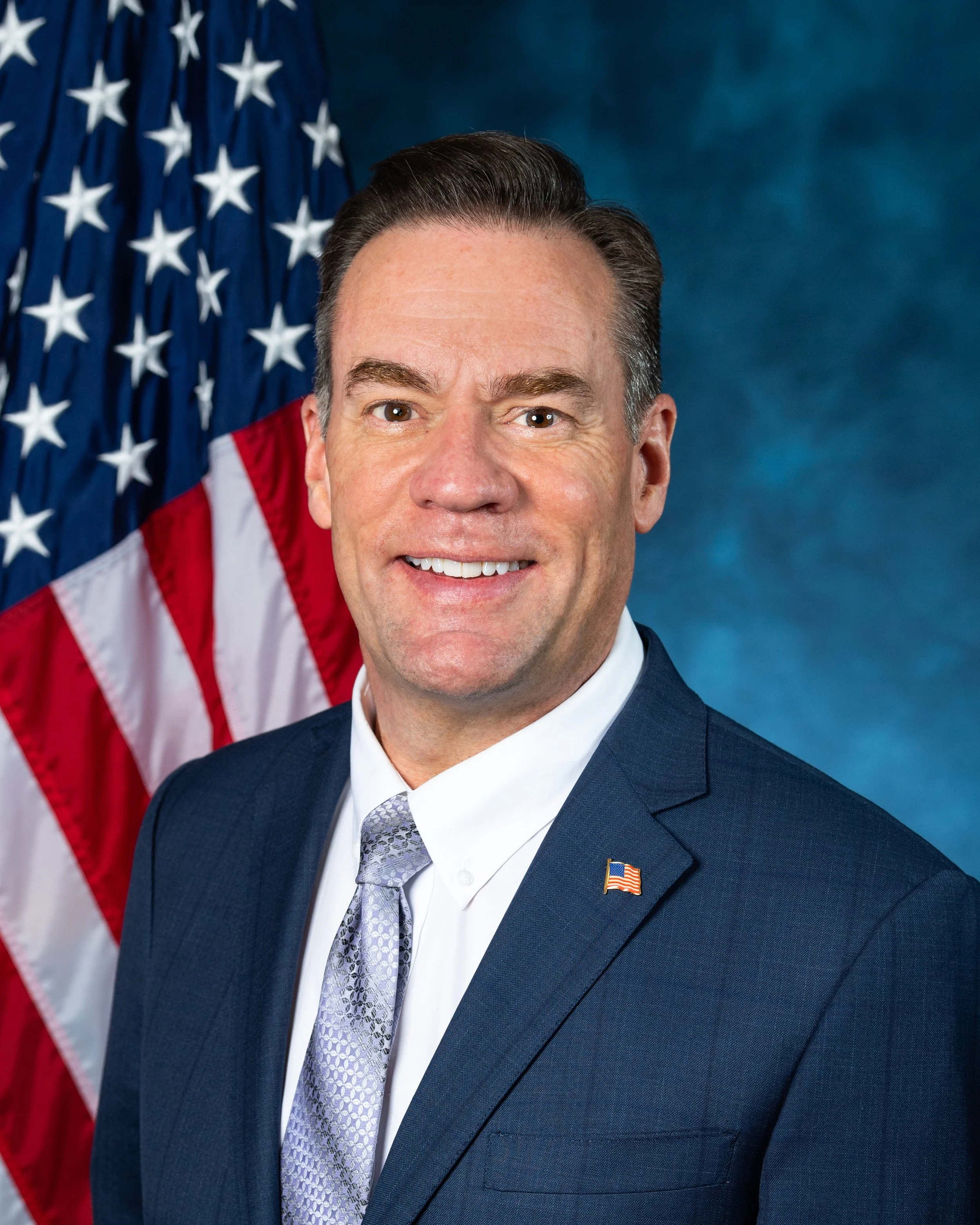 Close-up of a man in a suit, smiling, with American flag in the background.