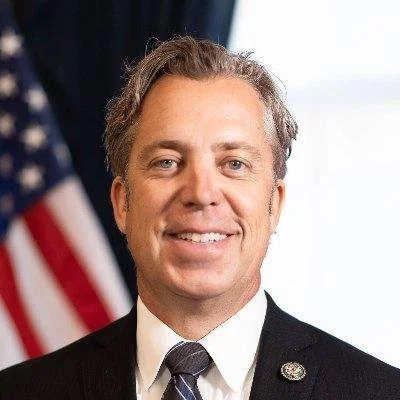 A portrait of a smiling man in a suit with an American flag in the background.