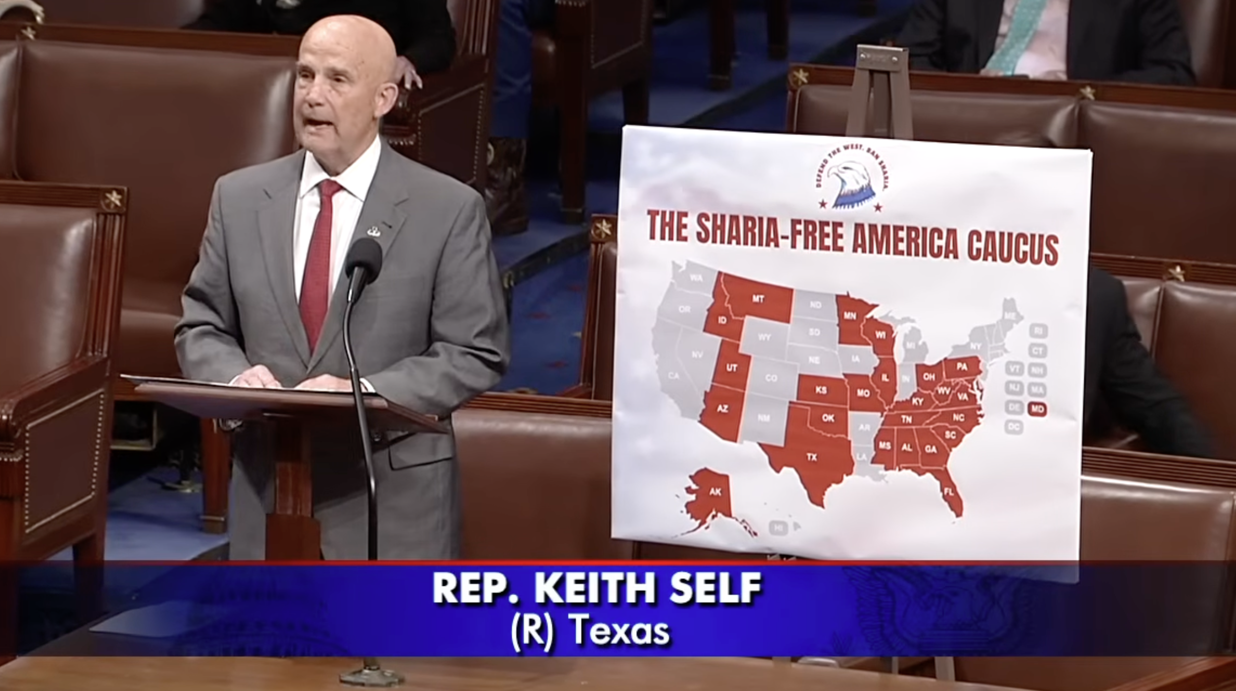 A man in a gray suit with a red tie standing at a podium with a microphone, speaking in a legislative chamber. Next to him is a large sign with a map of the United States highlighted with red states labeled 'The Sharia-Free America Caucus.' The sign is on an easel and features an eagle logo with the text 'Delivered to the West, and Beyond.' A portion of a news banner is visible at the bottom, showing 'REP. KEITH SELF (R) Texas.'