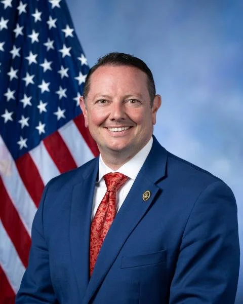 Man in a blue suit with a red patterned tie, smiling in front of an American flag on a cloudy blue background.