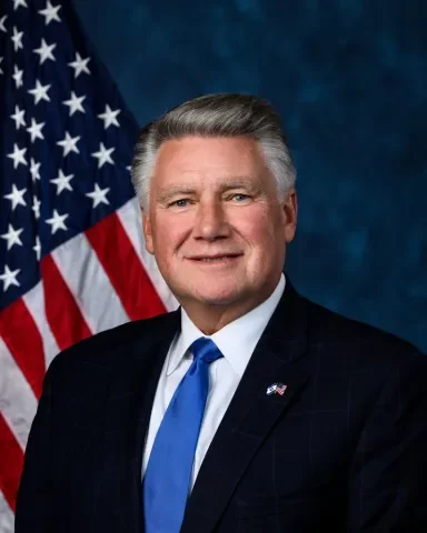 A formal portrait of a man in a dark suit, white shirt, and blue tie, standing in front of the American flag.