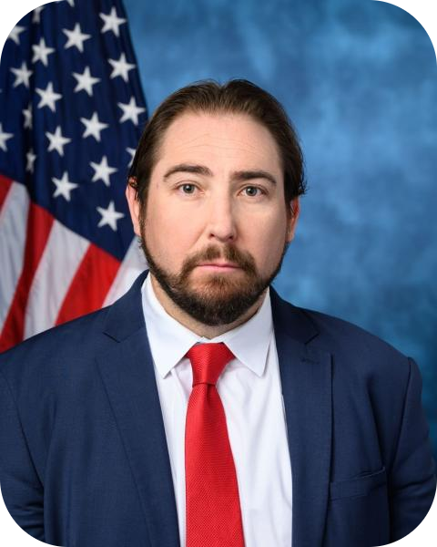 A man with dark hair and a beard wearing a navy blazer, white shirt, and red tie, posing in front of an American flag with a blue background.