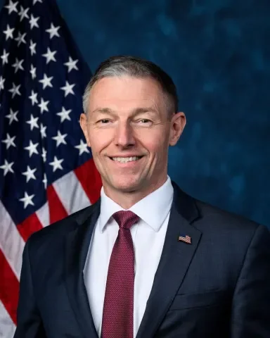 Professional portrait of a man in a suit standing in front of an American flag.