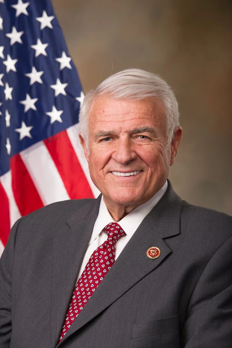 A man with white hair wearing a dark suit, white shirt, and maroon tie with white and blue dots, smiling, standing in front of an American flag.