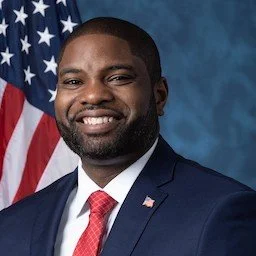 A smiling African American man in a navy suit, white shirt, and red tie, standing in front of an American flag.