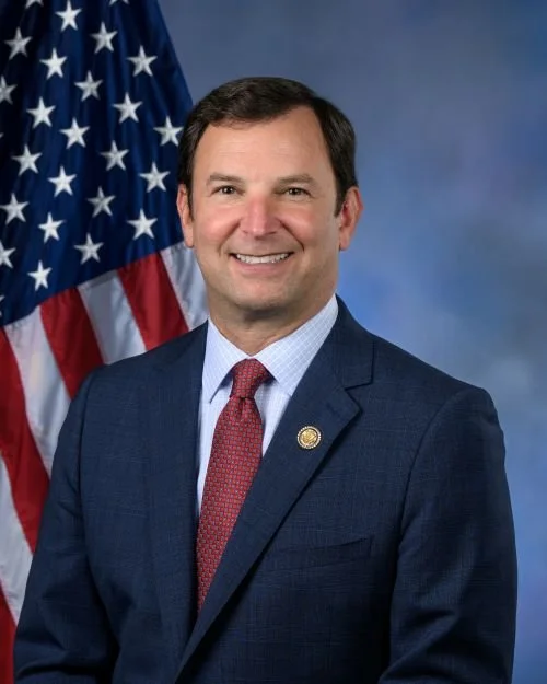 A man in a blue suit with a red tie standing in front of an American flag.