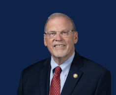 A middle-aged man with glasses, light skin, and short gray hair, wearing a dark suit, light blue shirt, and red patterned tie, smiling against a solid dark blue background.